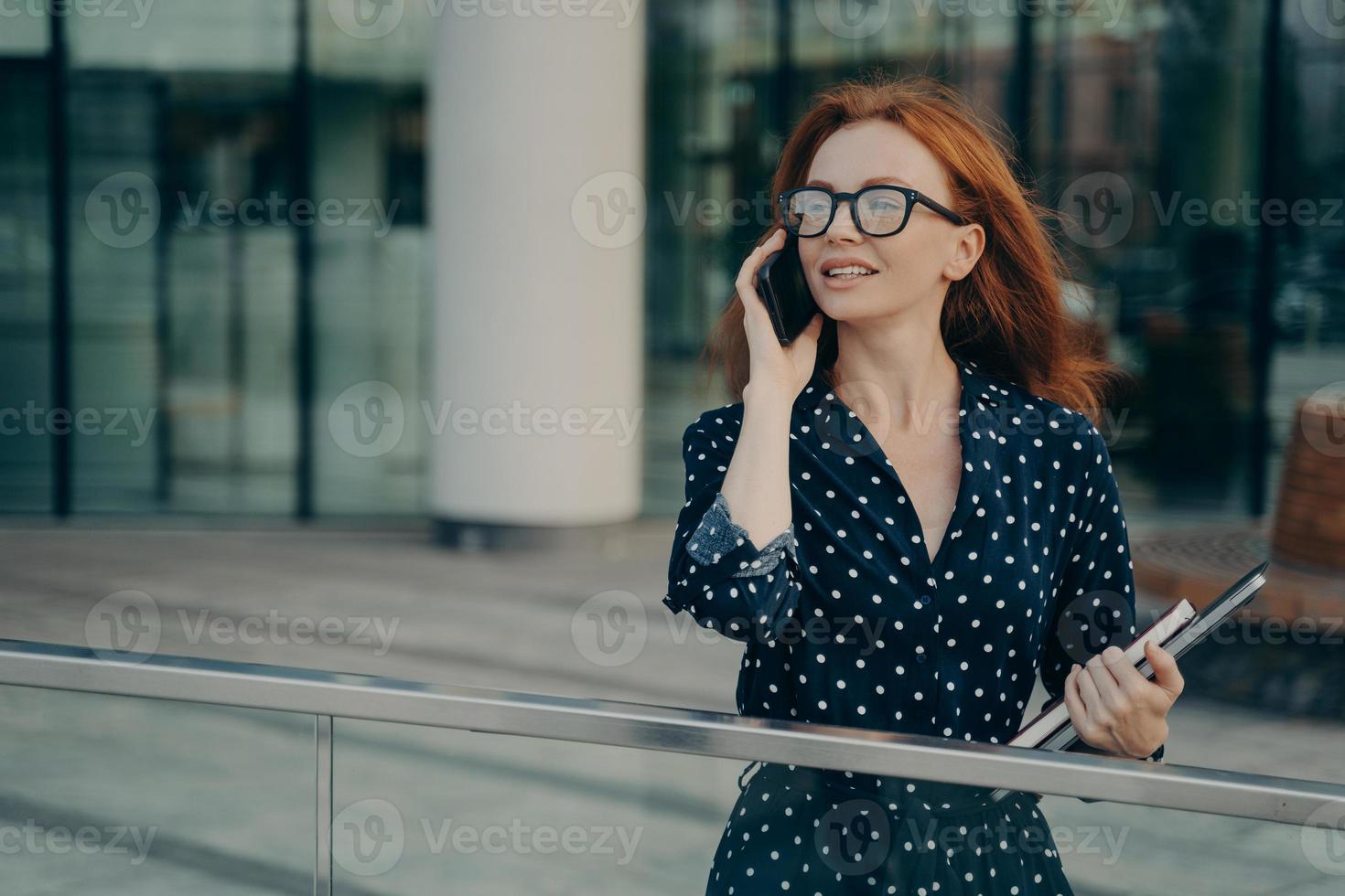 pelirroja pensativa mujer lleva gafas vestido de lunares tiene conversación telefónica foto