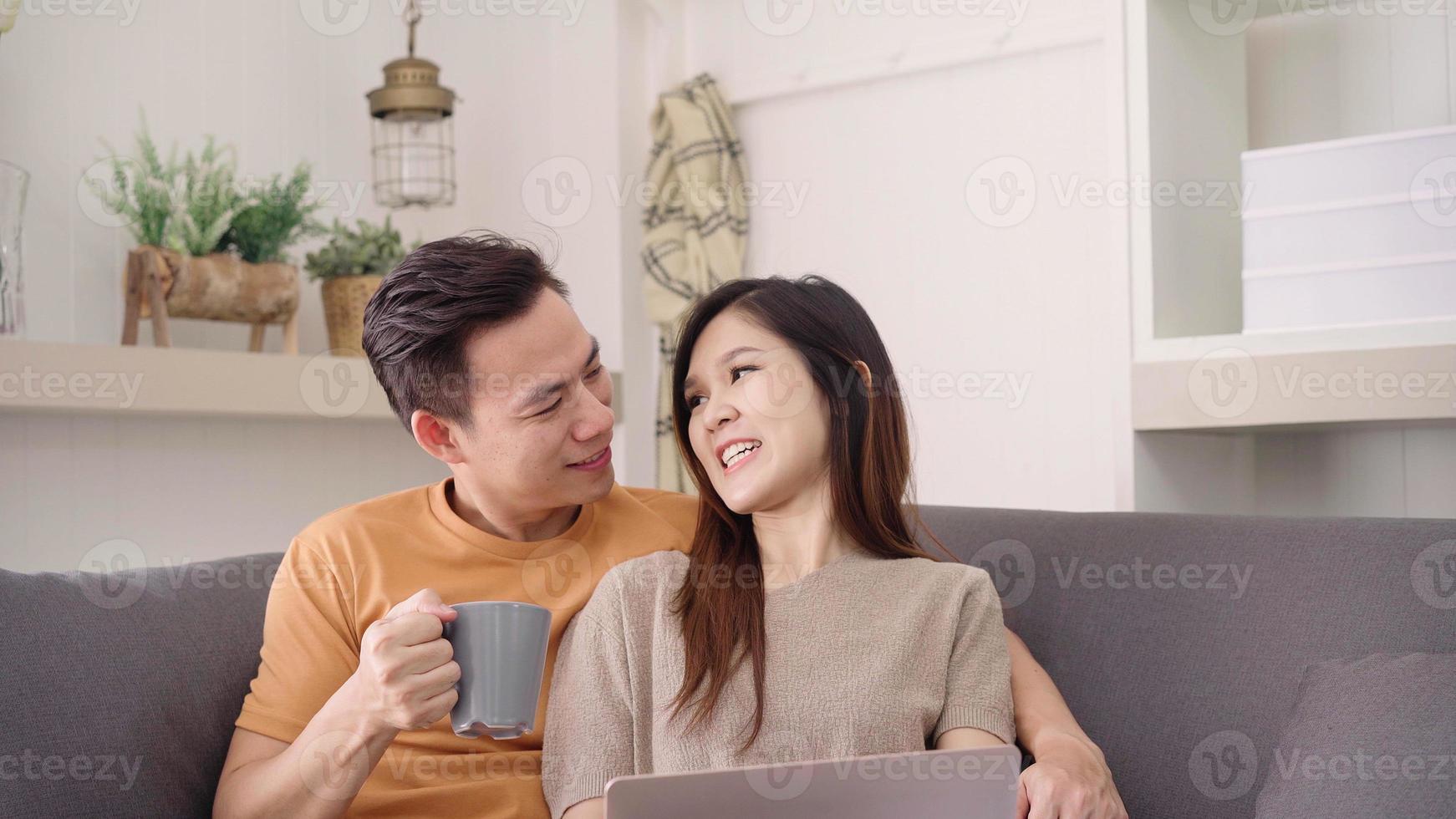 pareja asiática usando una laptop y tomando una taza de café caliente en la sala de estar en casa, una pareja dulce disfruta del momento de amor mientras se acuesta en el sofá cuando se relaja en casa. pareja de estilo de vida relajarse en el concepto de casa. foto