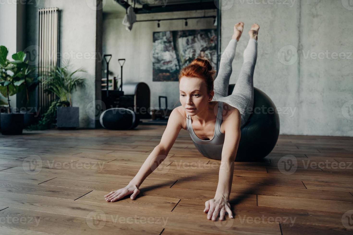 Athletic slender lady with red hair performing streching exercises on big silver fitball during pilates workout photo