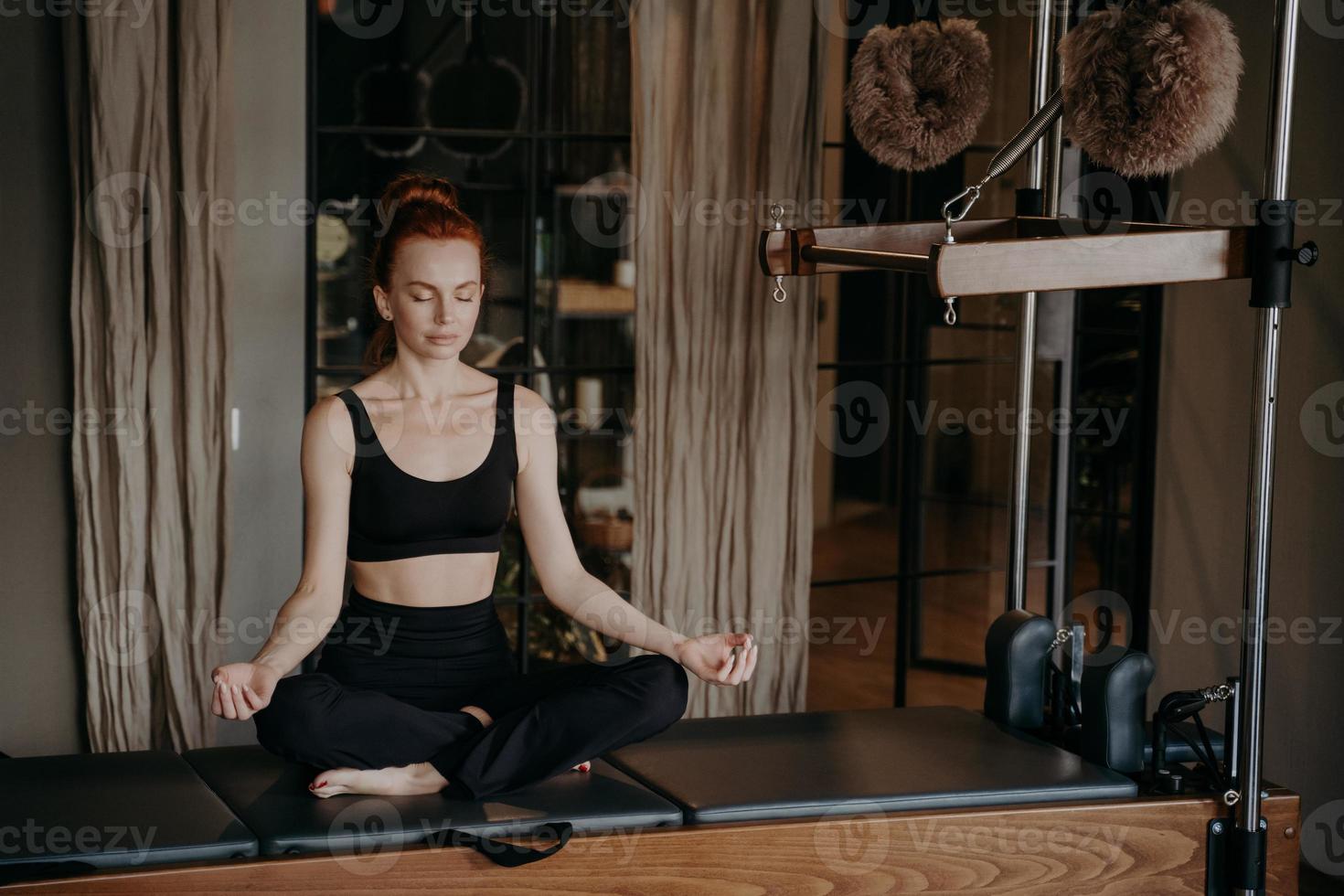 Young redhead woman in sportswear sitting in lotus position on trapeze table trying to relax photo