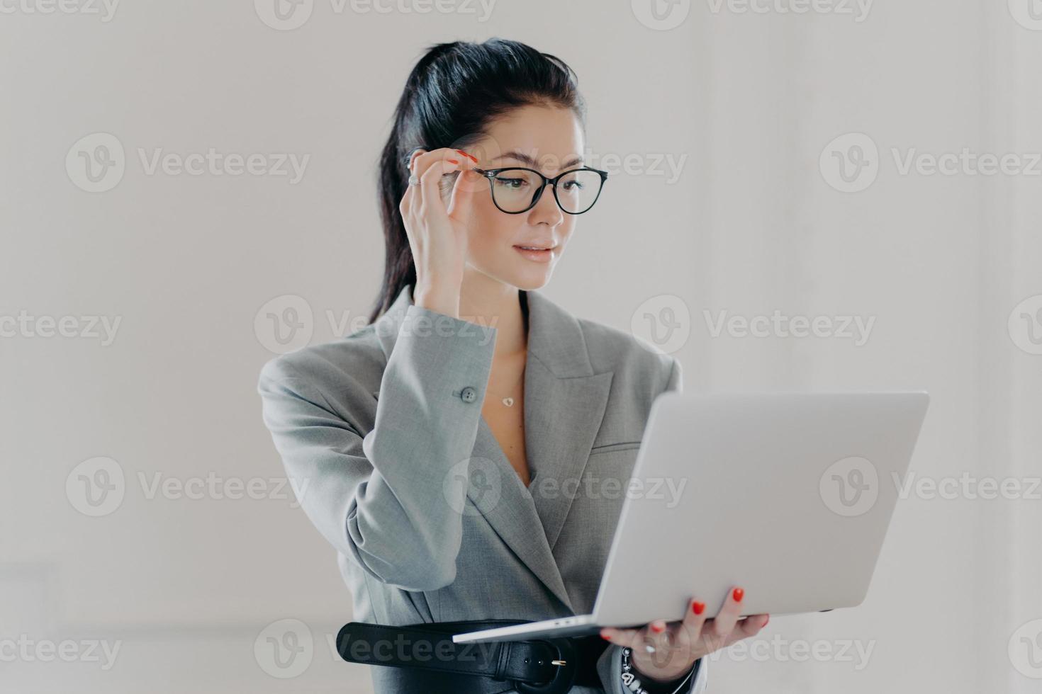 Photo of serious executive worker concentrated in laptop screen, wears transparent glasses for vision correction, browses internet web page, dressed in elegant formal clothes stands against white wall