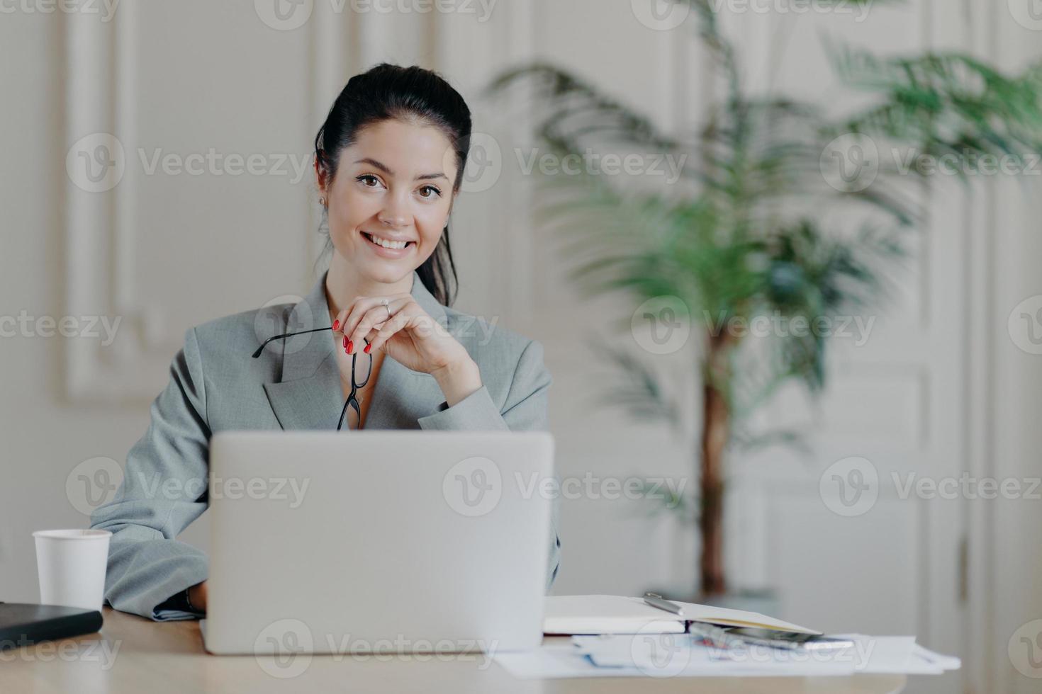 Charming stylish businesswoman works freelance at laptop computer, uses high speed internet connection, holds eyeglasses in hand, enjoys favorite remote job, reads publication, sits at office desk photo