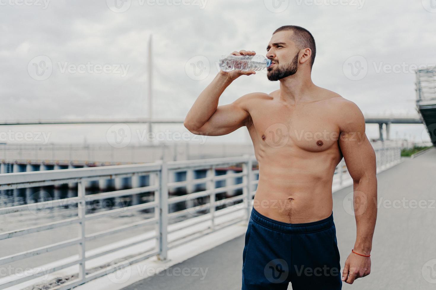 el deportista activo bebe una botella de agua posa con el torso desnudo concentrado en la distancia se siente sediento después de correr al aire libre lleno de energía, lleva un estilo de vida activo. deporte para una vida mejor foto