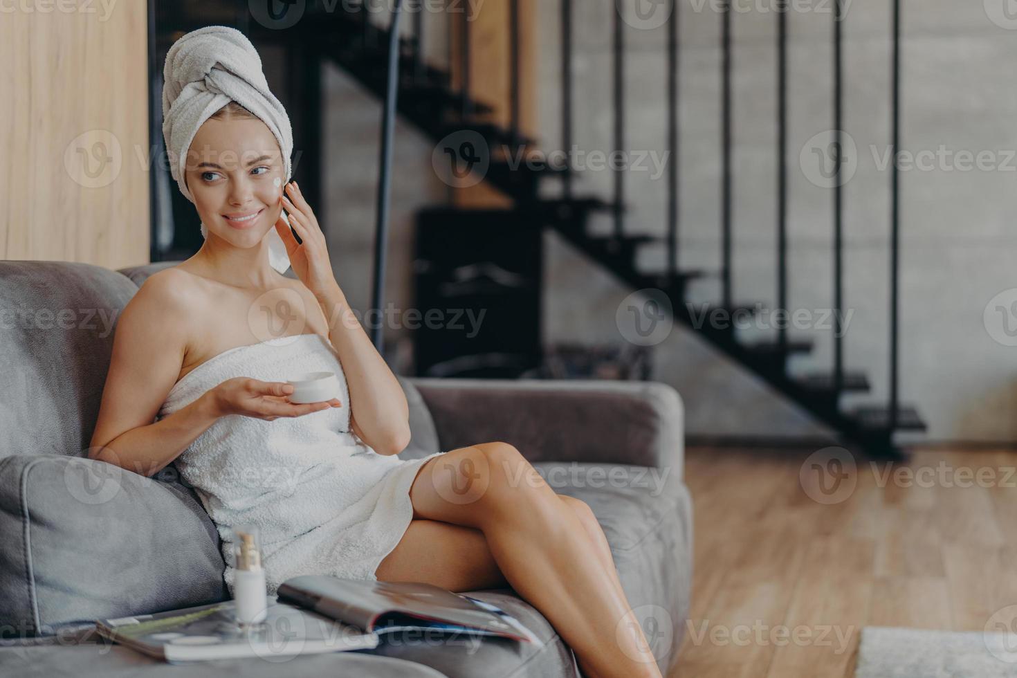 Pretty smiling European woman applies face cream on face, holds jar of cosmetic product, takes care of skin and complexion, poses on comfortable sofa with magazine against stairs background. photo
