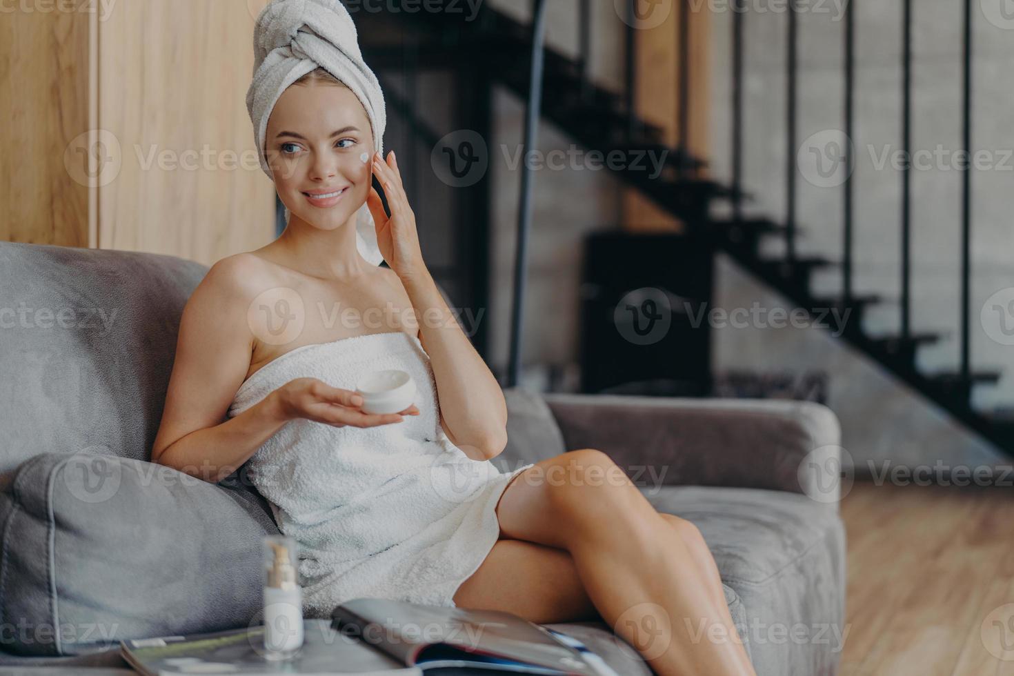 una mujer alegre de apariencia europea aplica crema facial para una piel suave y saludable, se sienta en un cómodo sofá, rodeada de una botella de loción y una revista, mira a un lado, tiene una complexión suave foto