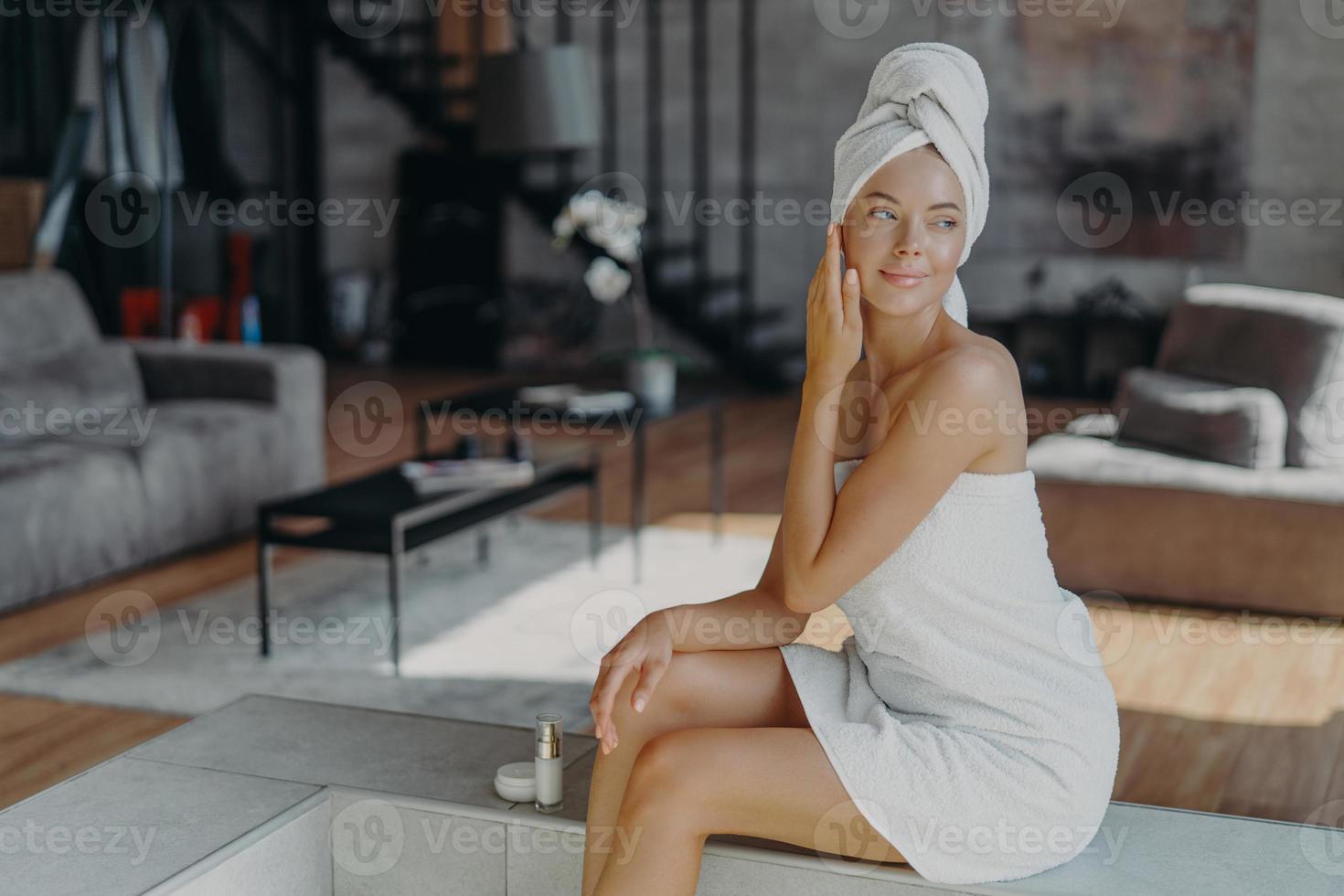Women, cosmetology, beauty and hygiene concept. Relaxed thoughtful woman wrapped in bath towel, applies anti wrinkle cream or body lotion, poses in living room at home, looks pensively right photo