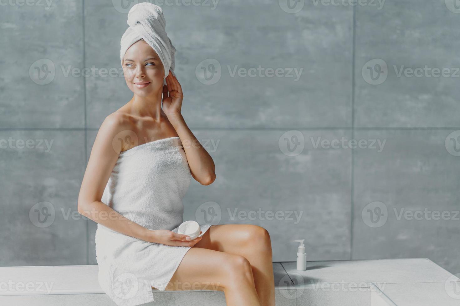 Photo of attractive thoughtful young woman looks away with dreamy expression, puts cream on body after taking bath, has clean fresh skin, poses with bare shoulders, enjoys taking spa procedures