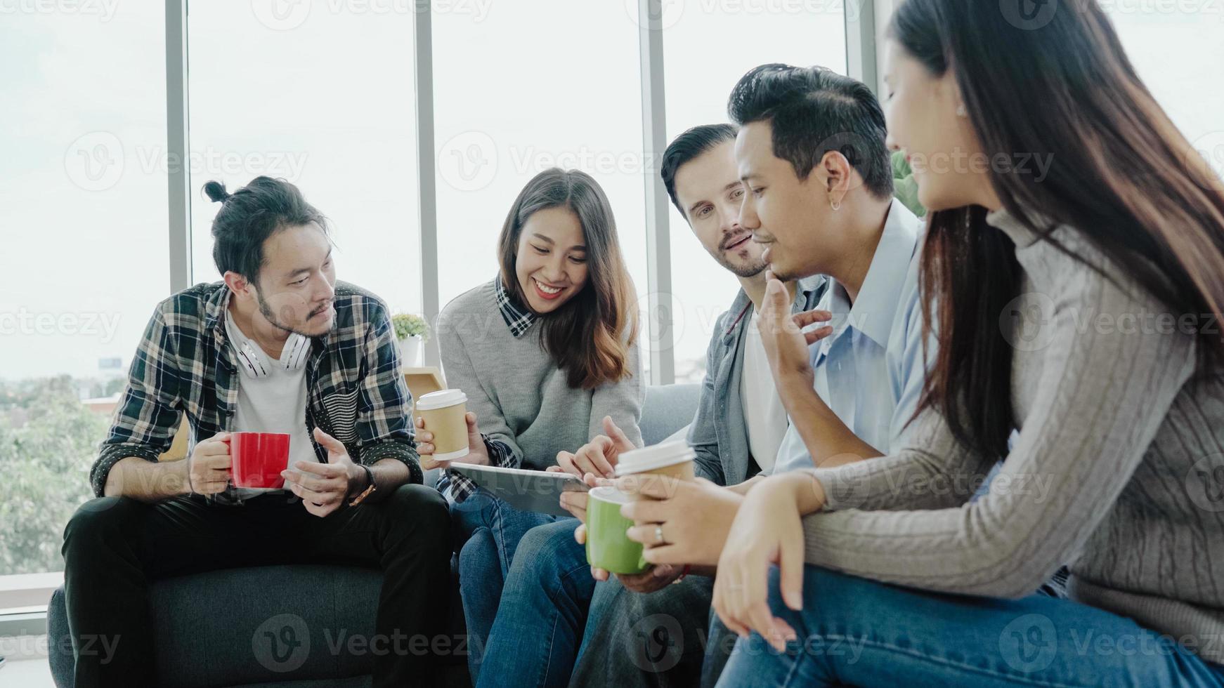 Multiethnic creative team diversity of young people group team holding coffee cups and discussing ideas meeting with tablet sitting on the couch at office. Coffee break time at creative office. photo