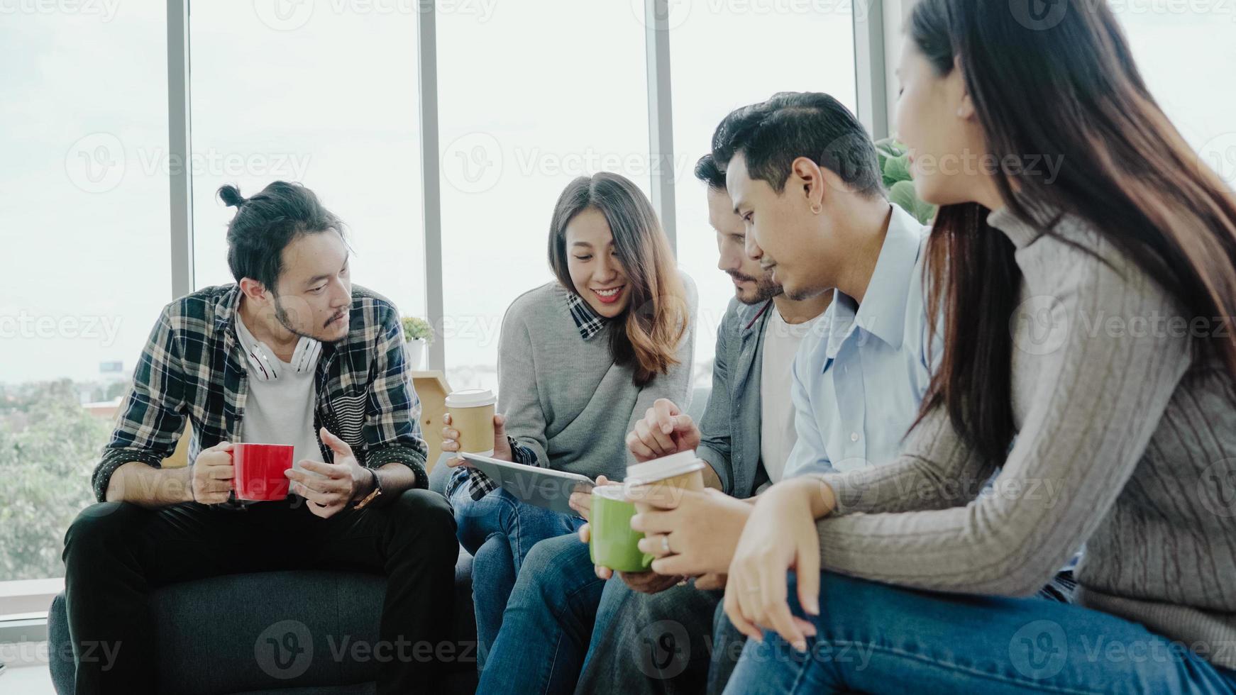 Multiethnic creative team diversity of young people group team holding coffee cups and discussing ideas meeting with tablet sitting on the couch at office. Coffee break time at creative office. photo