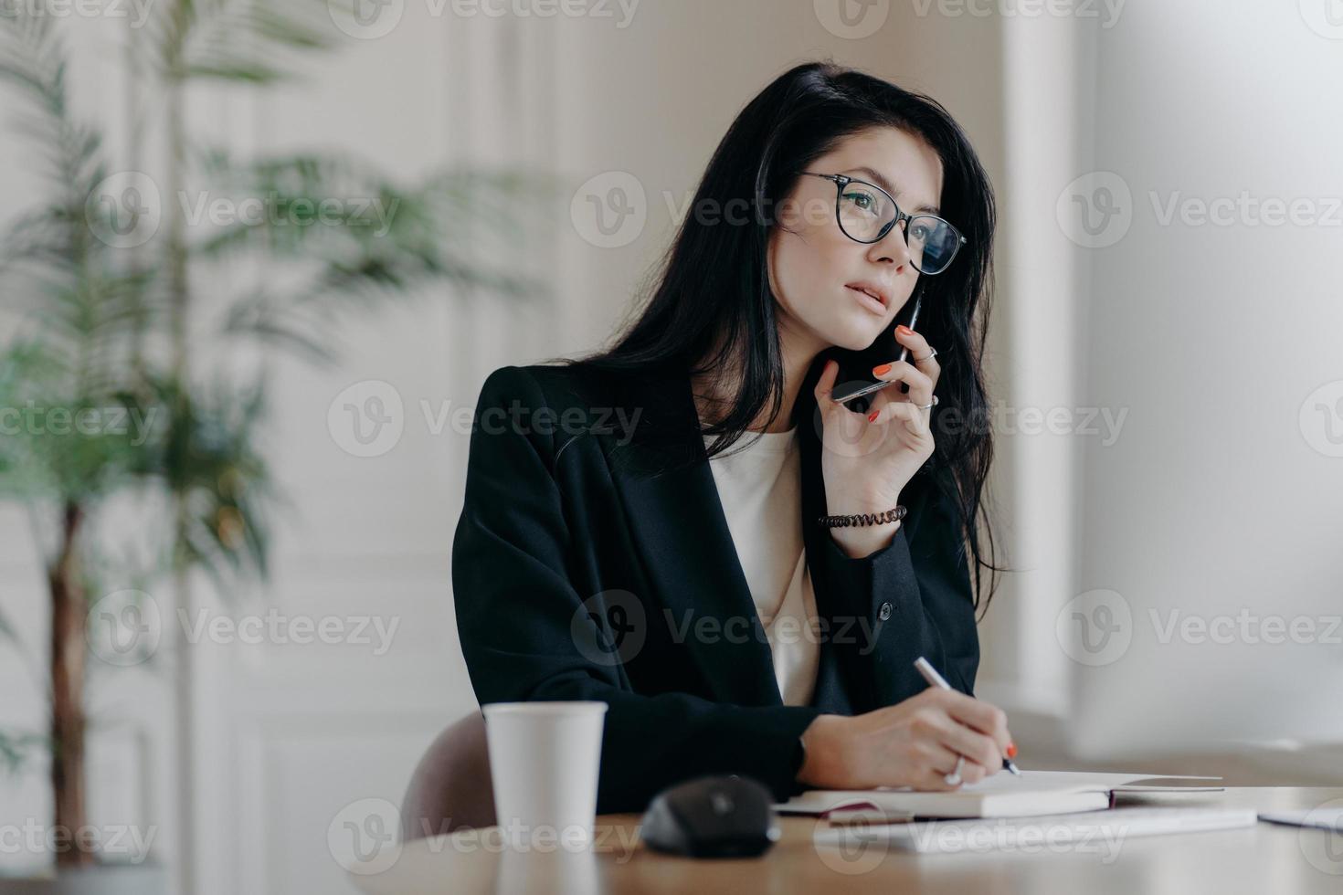 Gorgeous businesswoman makes phone call, concentrated in computer screen, writes down information into notepad, poses at cozy workplace, focused on important task, makes notes in personal organizer photo