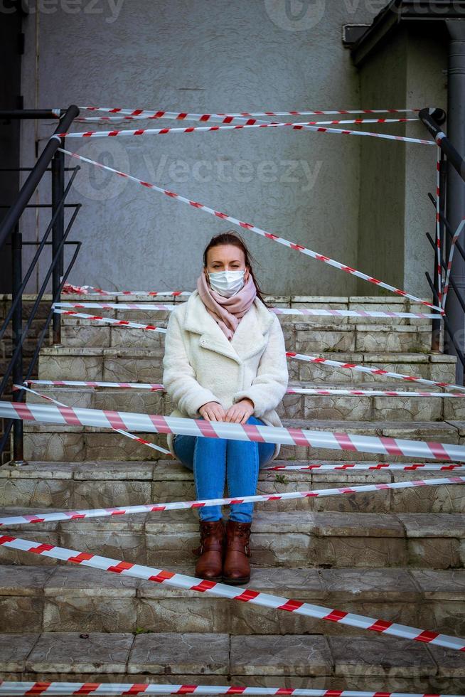 Photo of a girl in a mask. Sitting on the street with danger warning tapes.