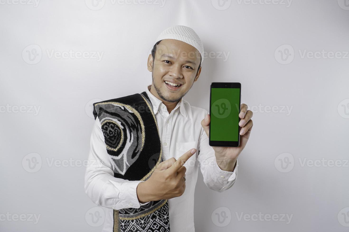 A portrait of a happy Asian Muslim man smiling while showing copy space on his phone, isolated by white background photo