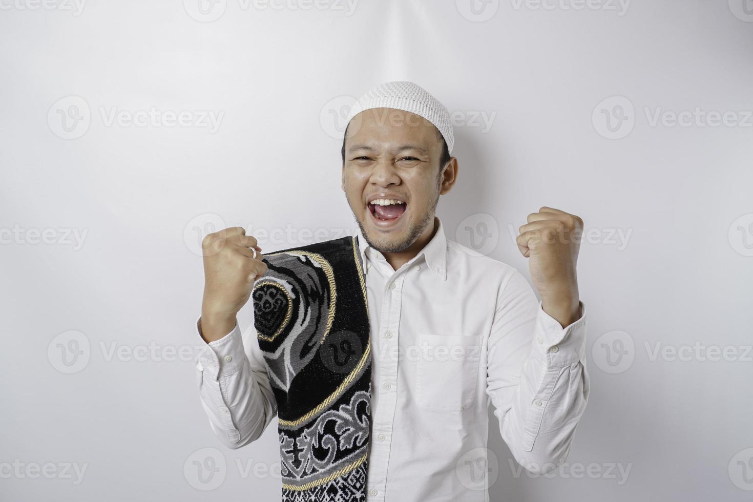 A happy young Asian Muslim man with a prayer rug on his shoulder showing a successful expression isolated by white background photo