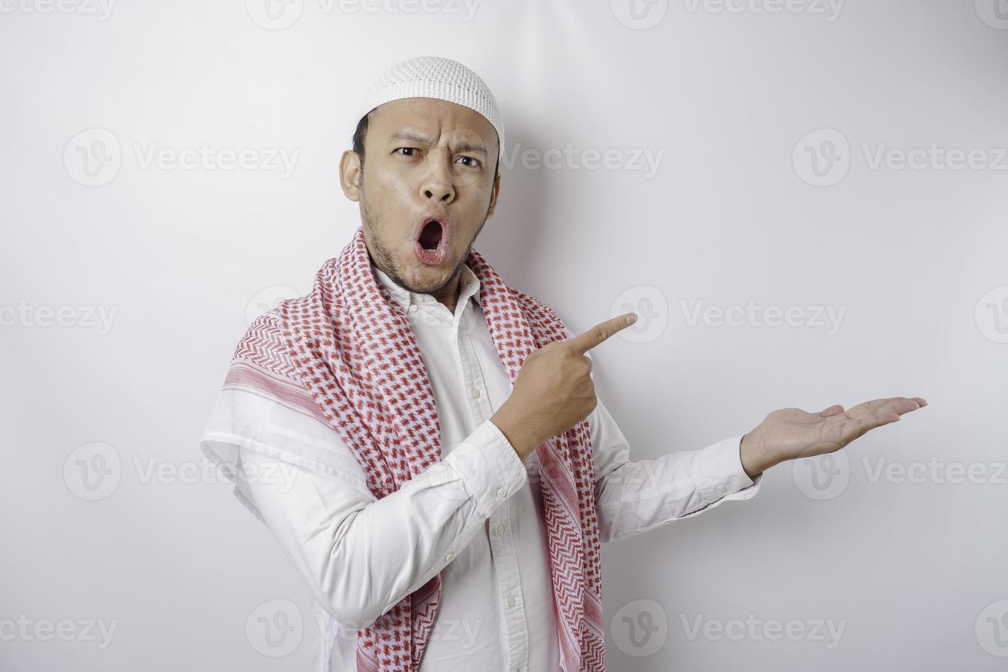 Shocked Asian Muslim man pointing at the copy space beside him, isolated by white background photo