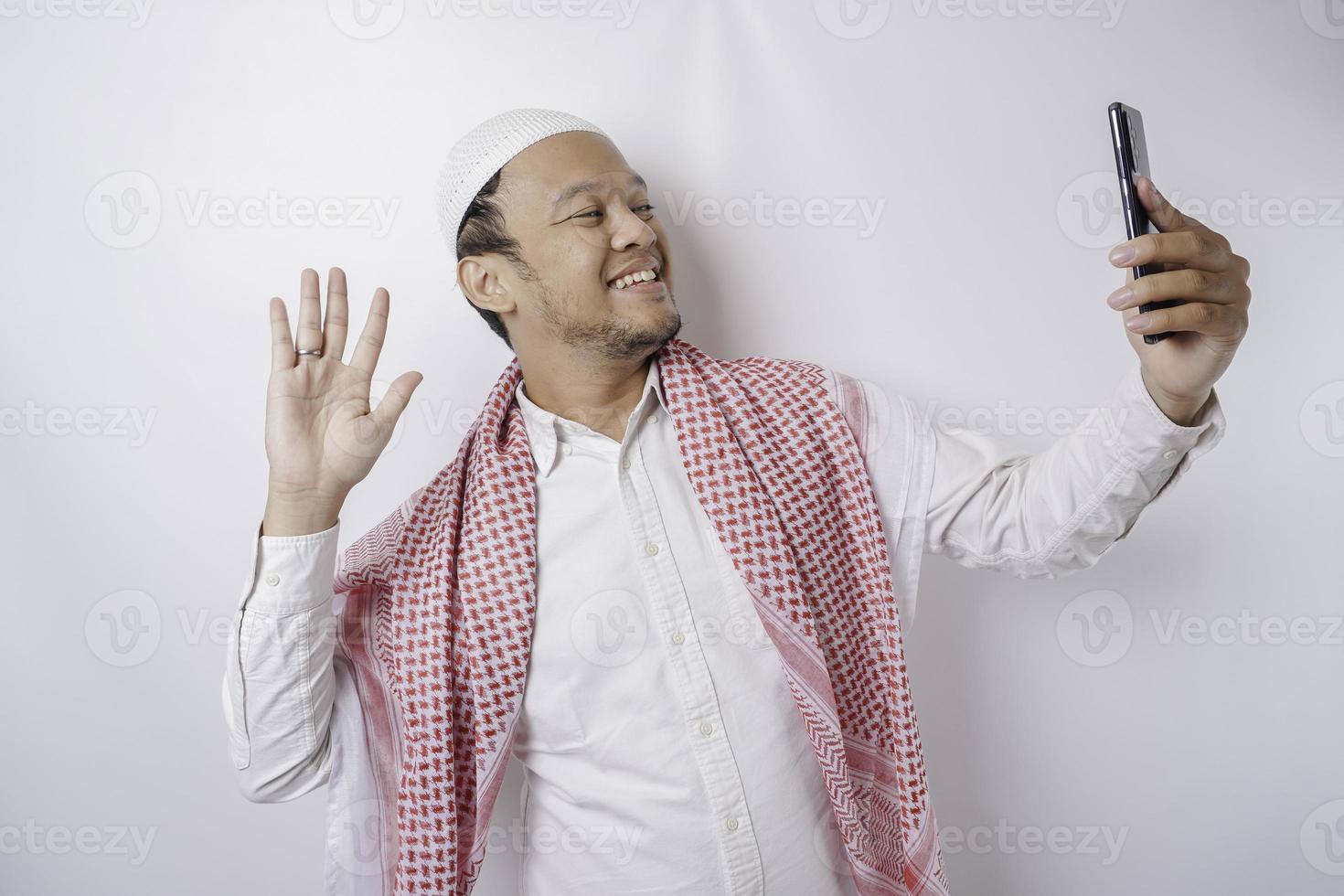 A portrait of a happy Asian Muslim man smiling while holding his phone, isolated by white background photo