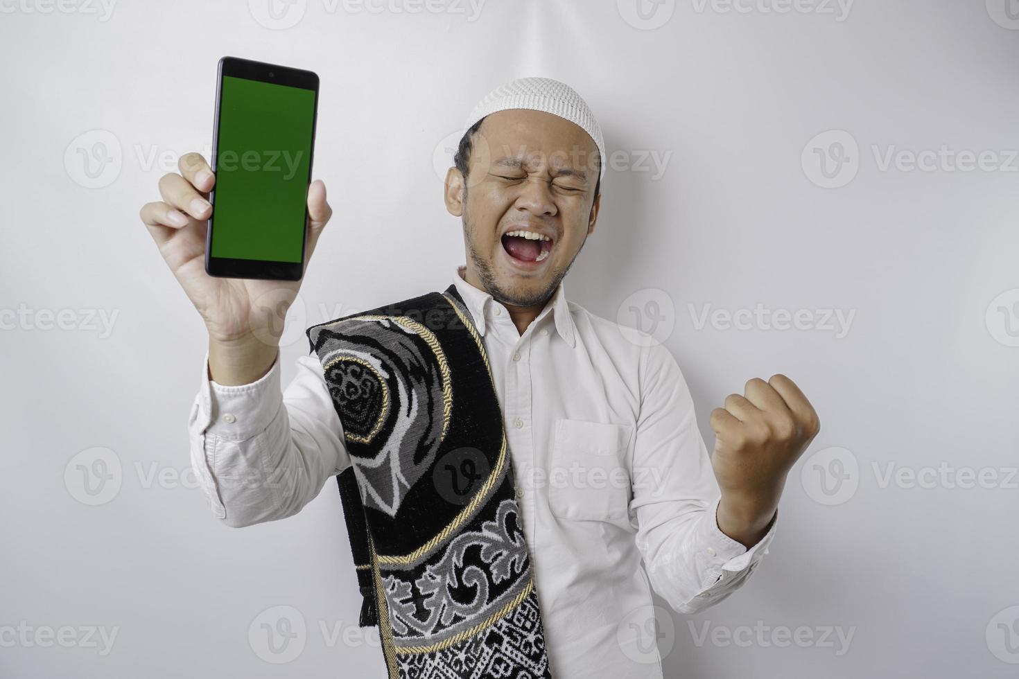 A happy young Asian Muslim man with a successful expression showing copy space on his phone isolated by white background photo