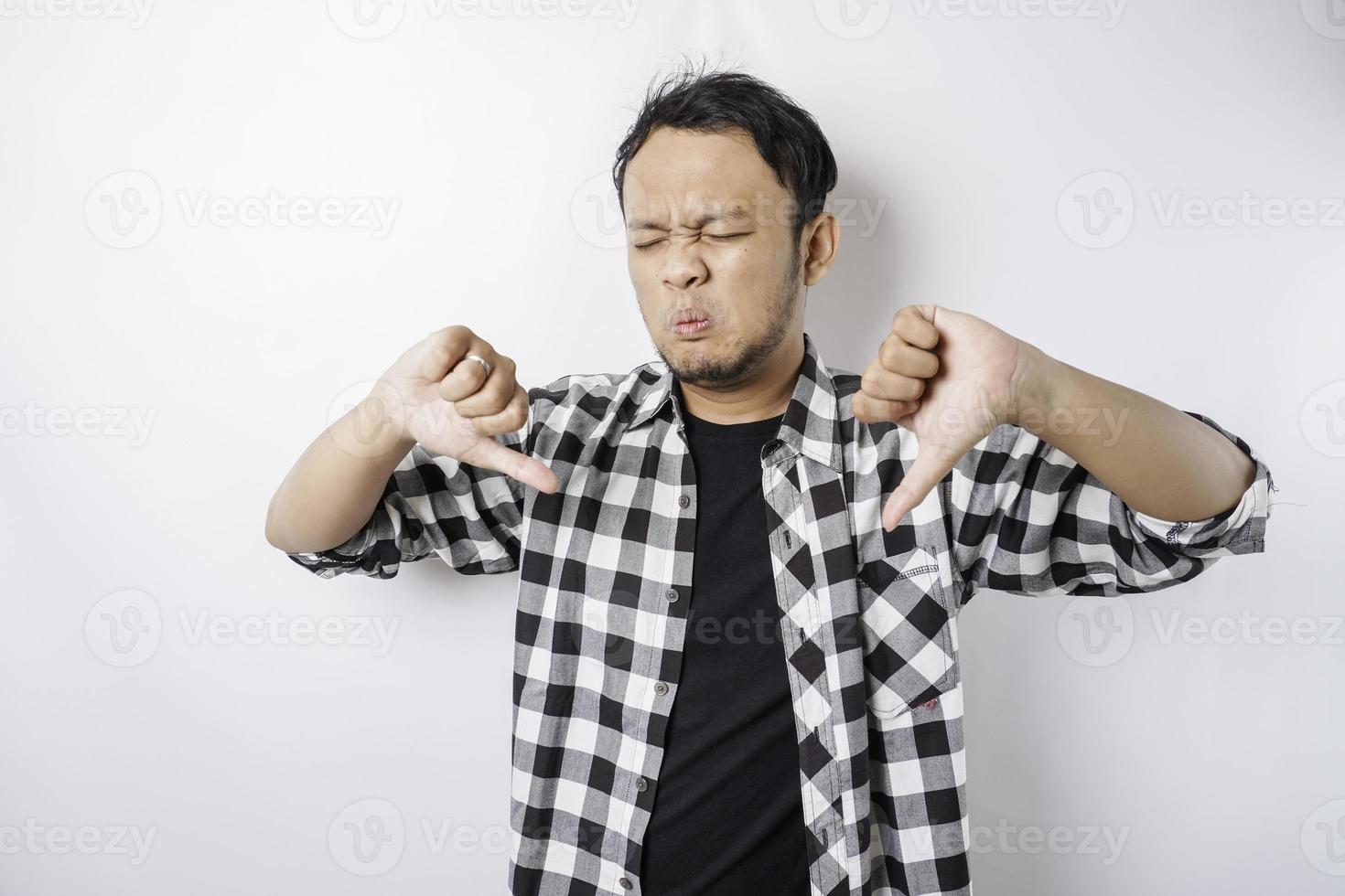 Disappointed Asian man wearing tartan short gives thumbs down hand gesture of approval, isolated by white background photo