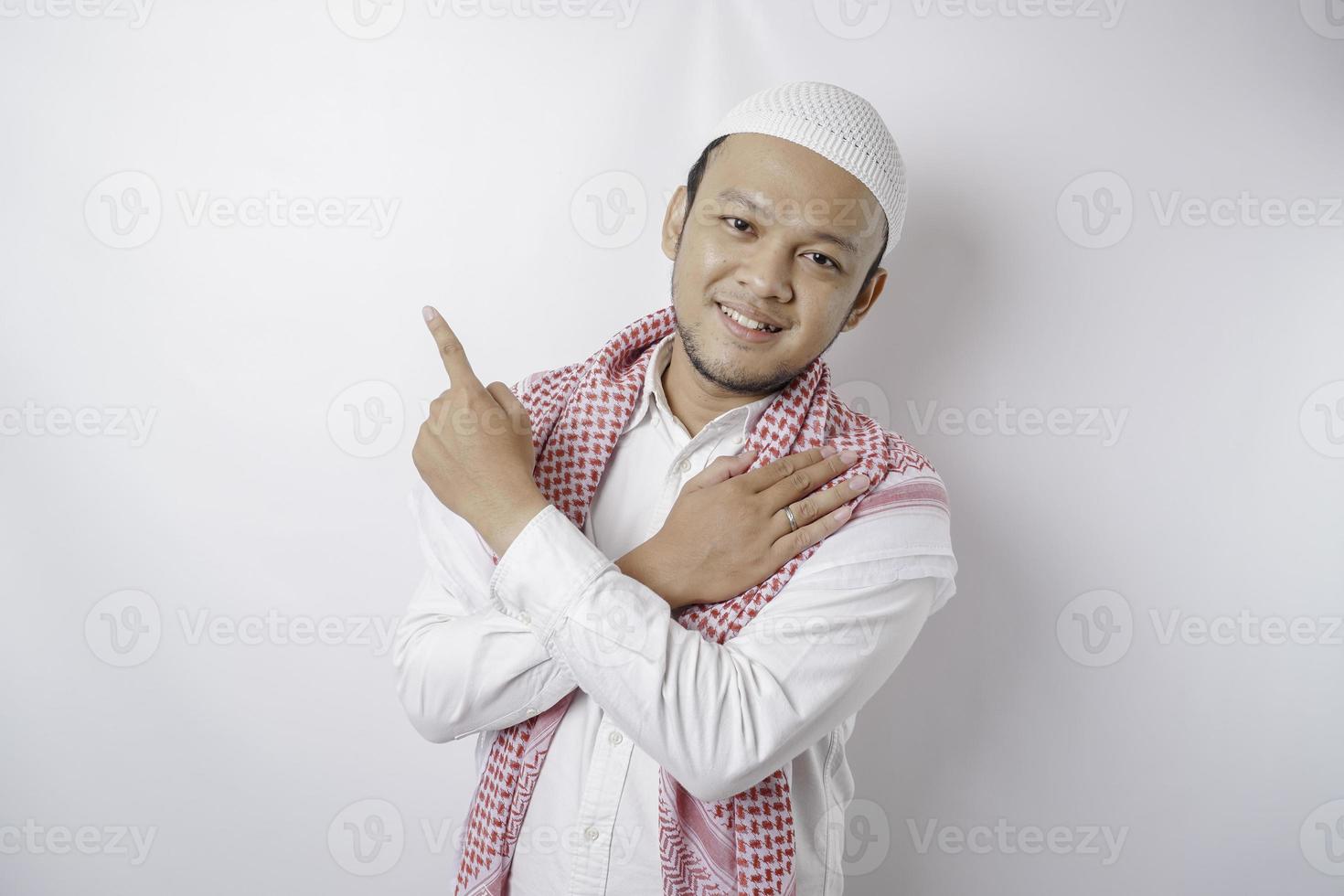 Smiling Asian Muslim man pointing at the copy space on top of him, isolated by white background photo