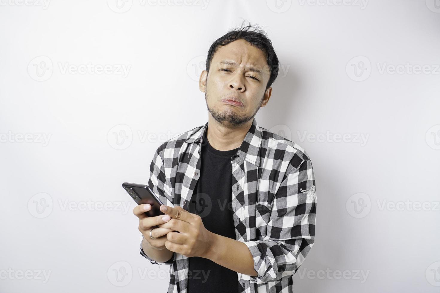 un joven asiático insatisfecho se ve descontento usando una camisa de tartán expresiones faciales irritadas sosteniendo su teléfono foto