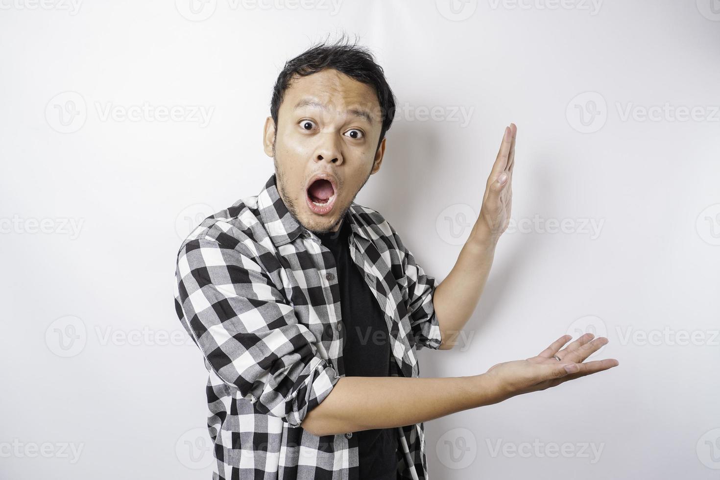 Shocked Asian man wearing tartan shirt is pointing at the copy space beside him, isolated by white background photo