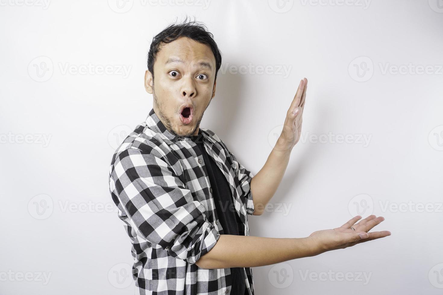 Shocked Asian man wearing tartan shirt is pointing at the copy space beside him, isolated by white background photo