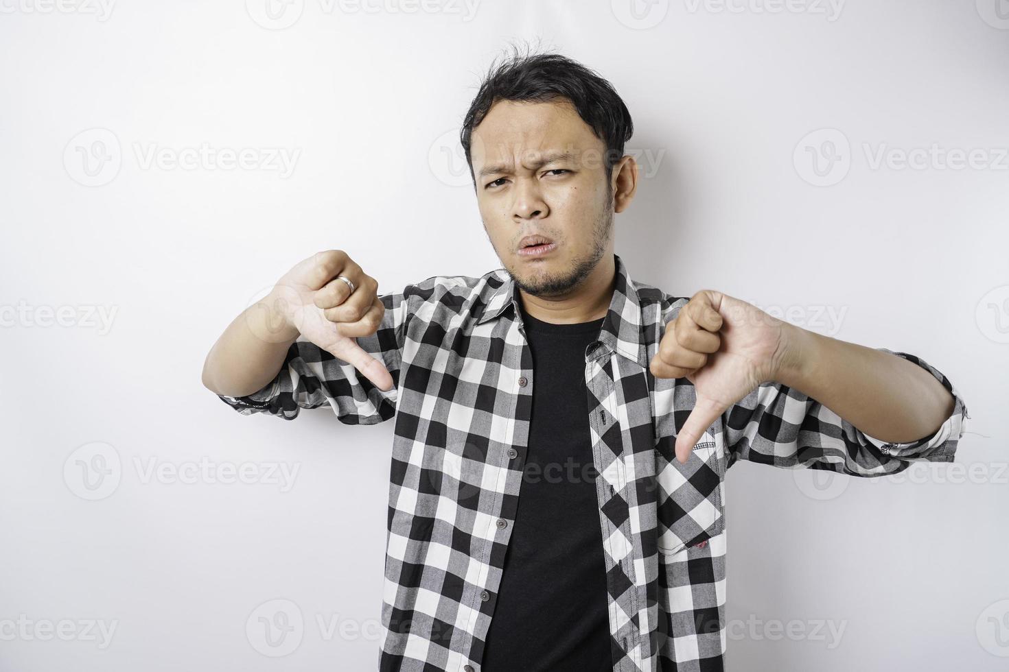 Disappointed Asian man wearing tartan short gives thumbs down hand gesture of approval, isolated by white background photo