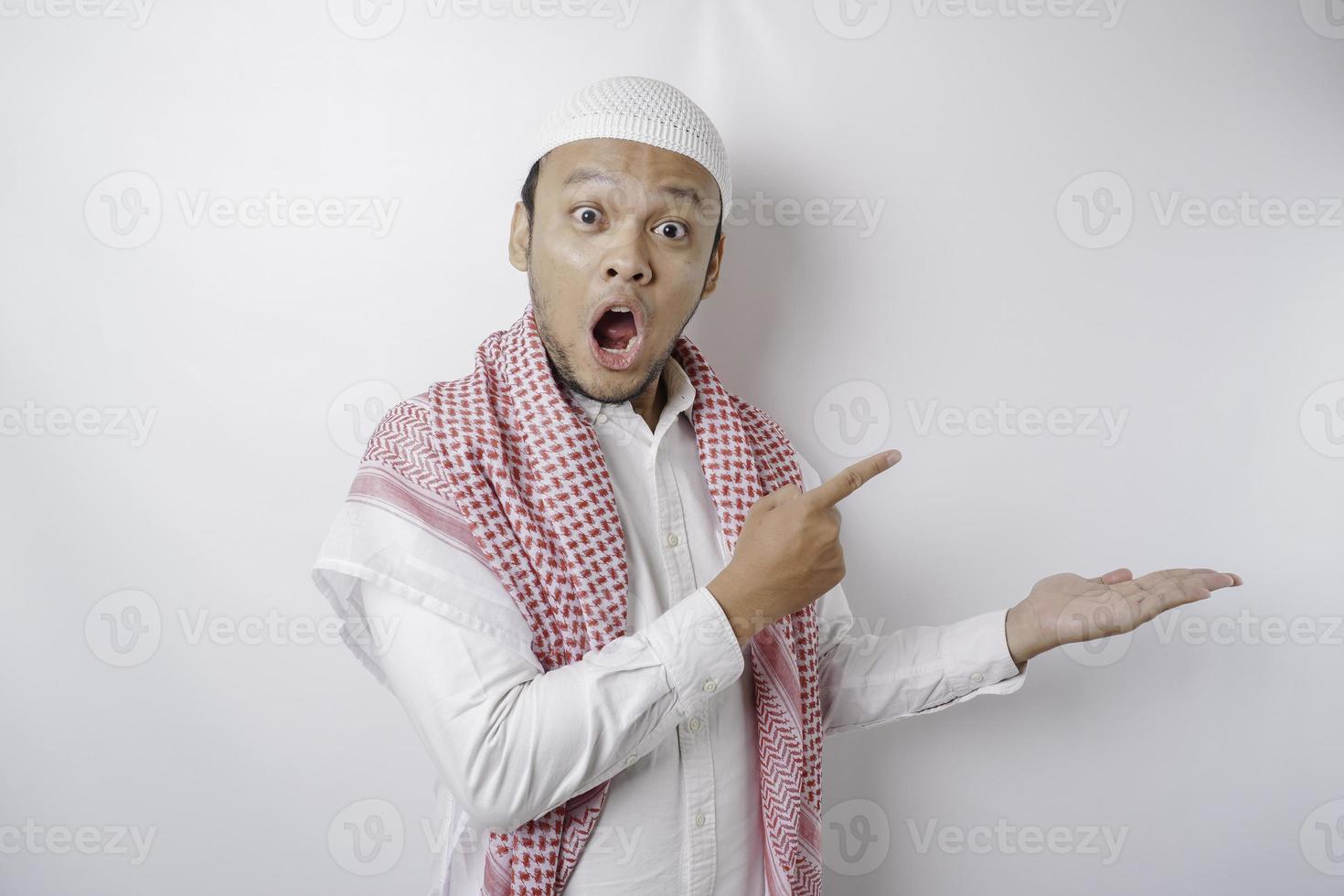 Shocked Asian Muslim man pointing at the copy space beside him, isolated by white background photo