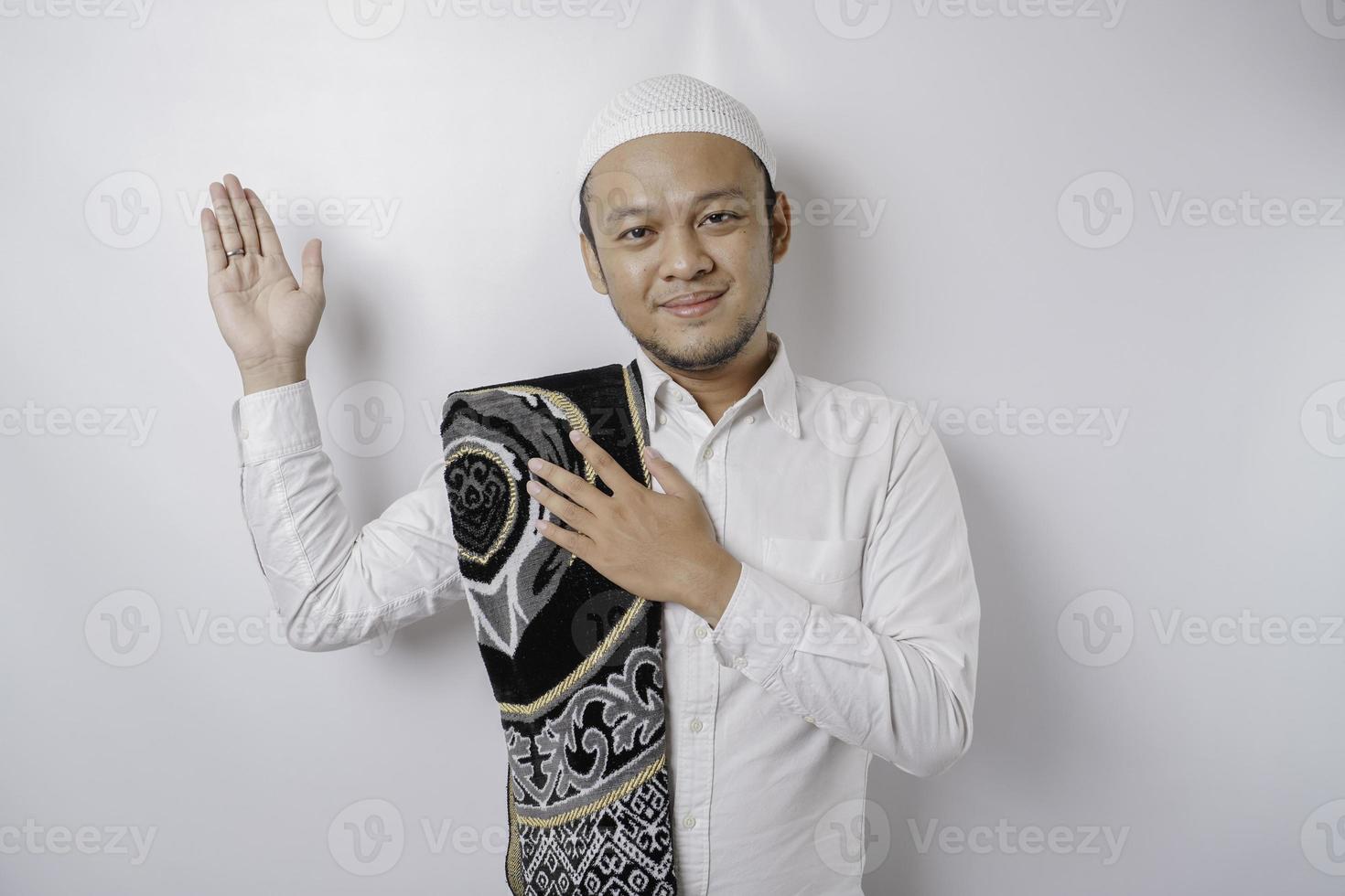 Smiling Asian Muslim man with prayer rug on his shoulder is pointing at the copy space on top of him, isolated by white background photo