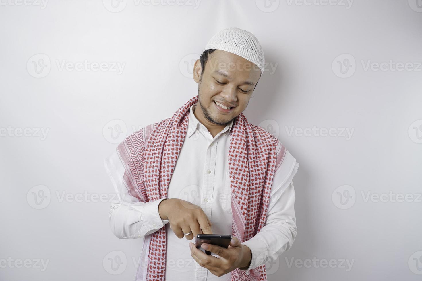 A portrait of a happy Asian Muslim man smiling while holding his phone, isolated by white background photo