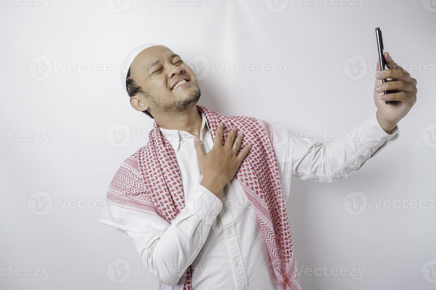 A portrait of a happy Asian Muslim man smiling while holding his phone, isolated by white background photo