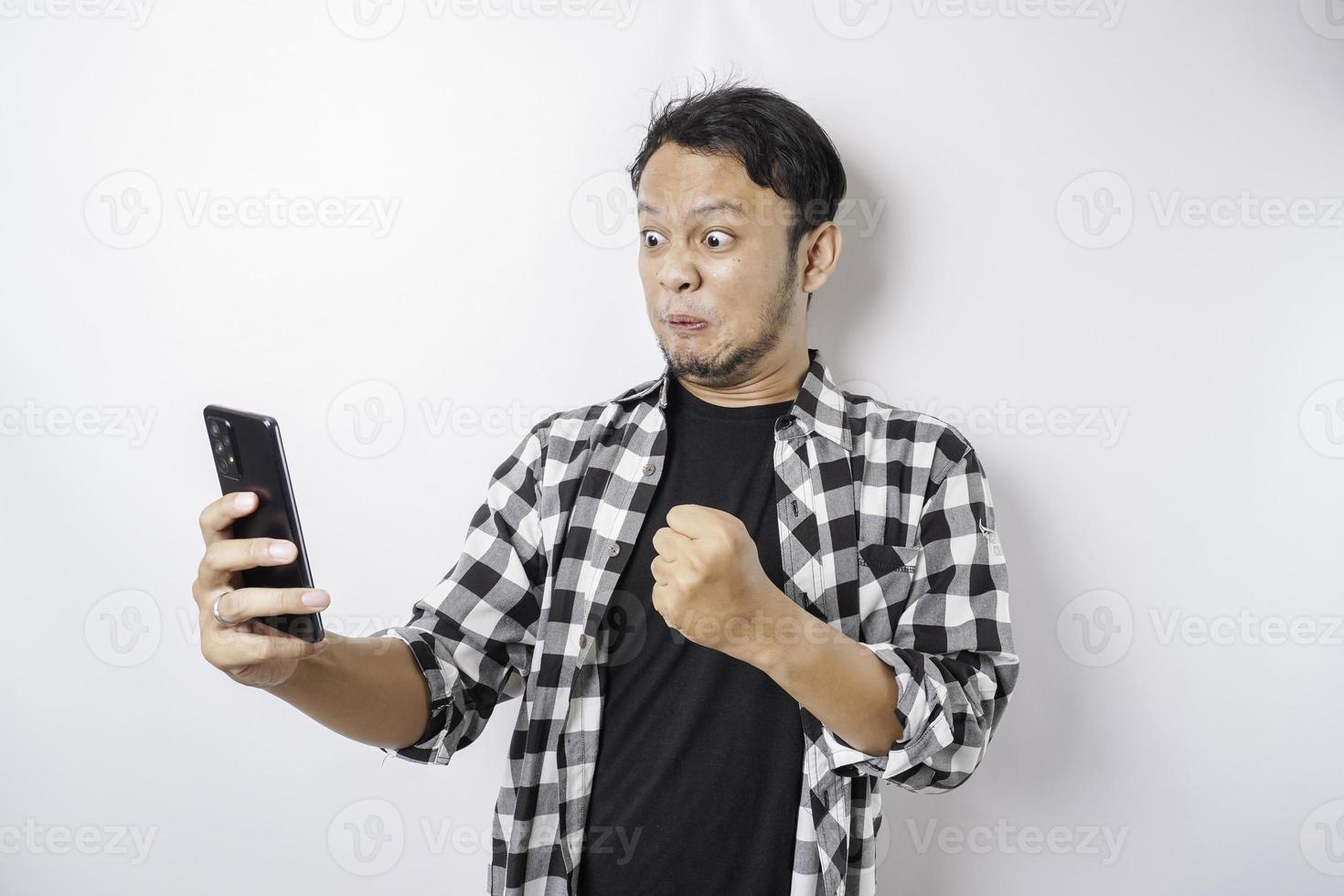 The angry and mad face of Asian man in tartan shirt while holding his phone on isolated white background. photo