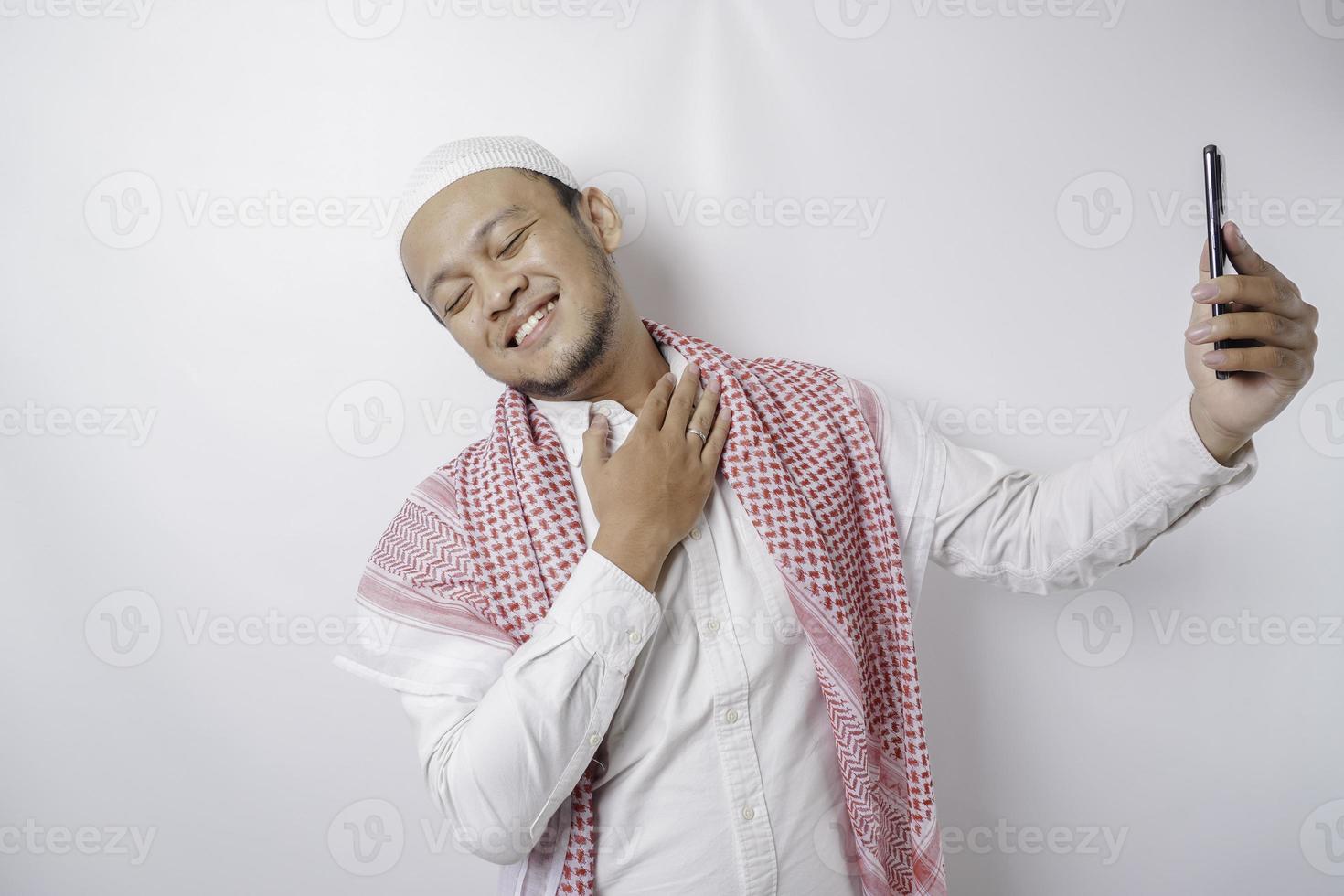 un retrato de un musulmán asiático feliz sonriendo mientras sostiene su teléfono, aislado por fondo blanco foto