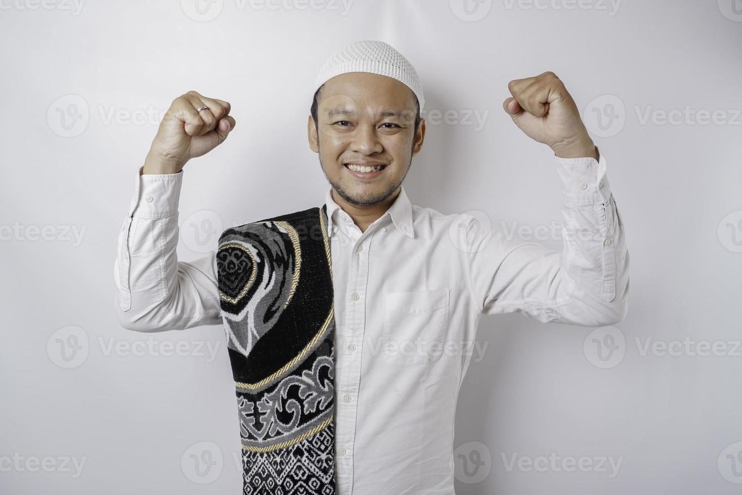 A happy young Asian Muslim man with a prayer rug on his shoulder showing a successful expression isolated by white background photo