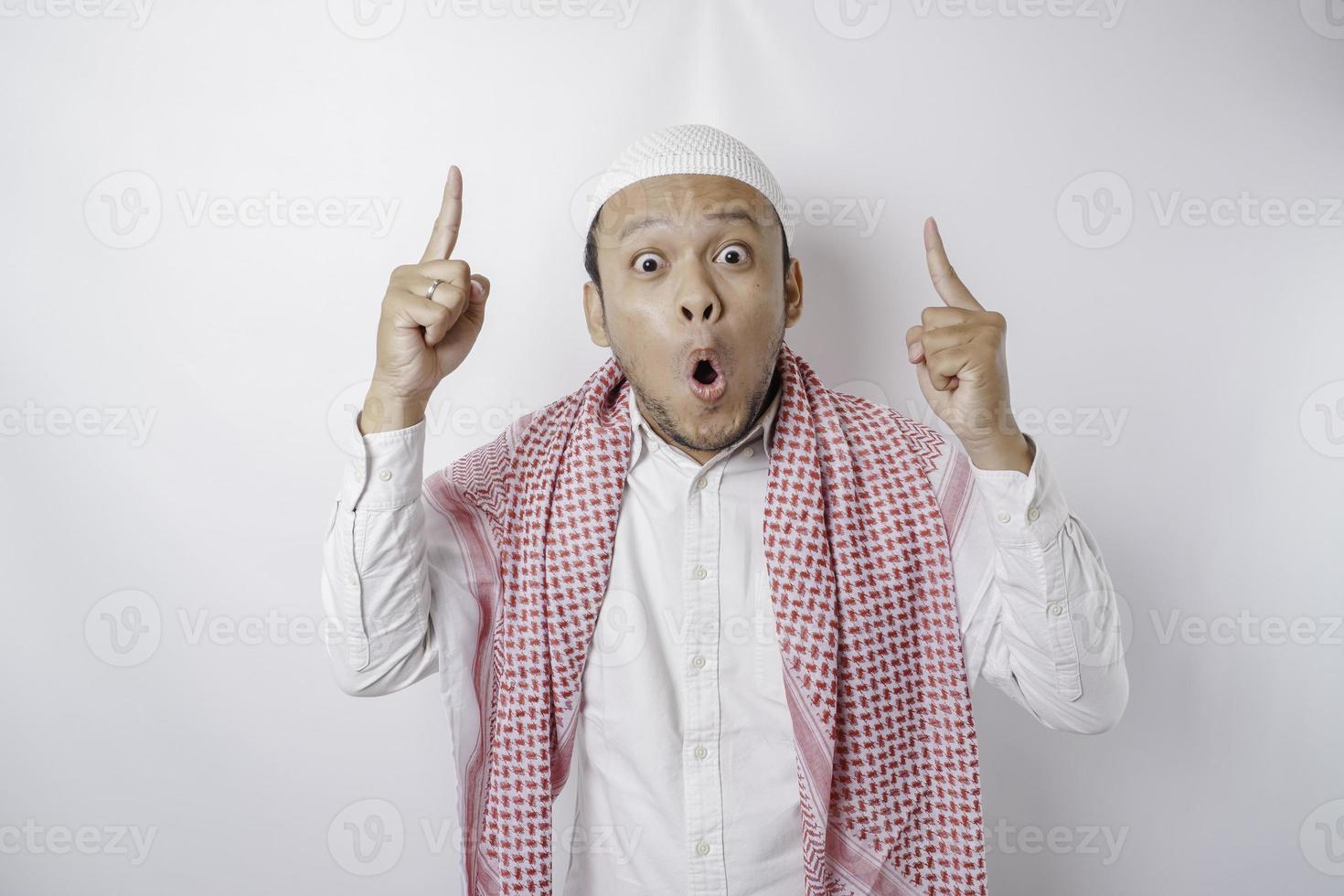 Shocked Asian Muslim man pointing at the copy space on top of him, isolated by a white background photo