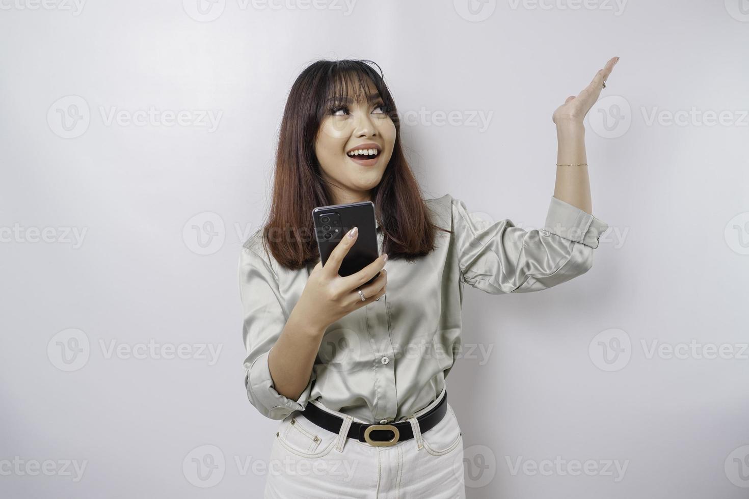 mujer asiática emocionada con traje verde salvia apuntando al espacio de copia encima de ella mientras sostiene su teléfono, aislada por fondo blanco foto