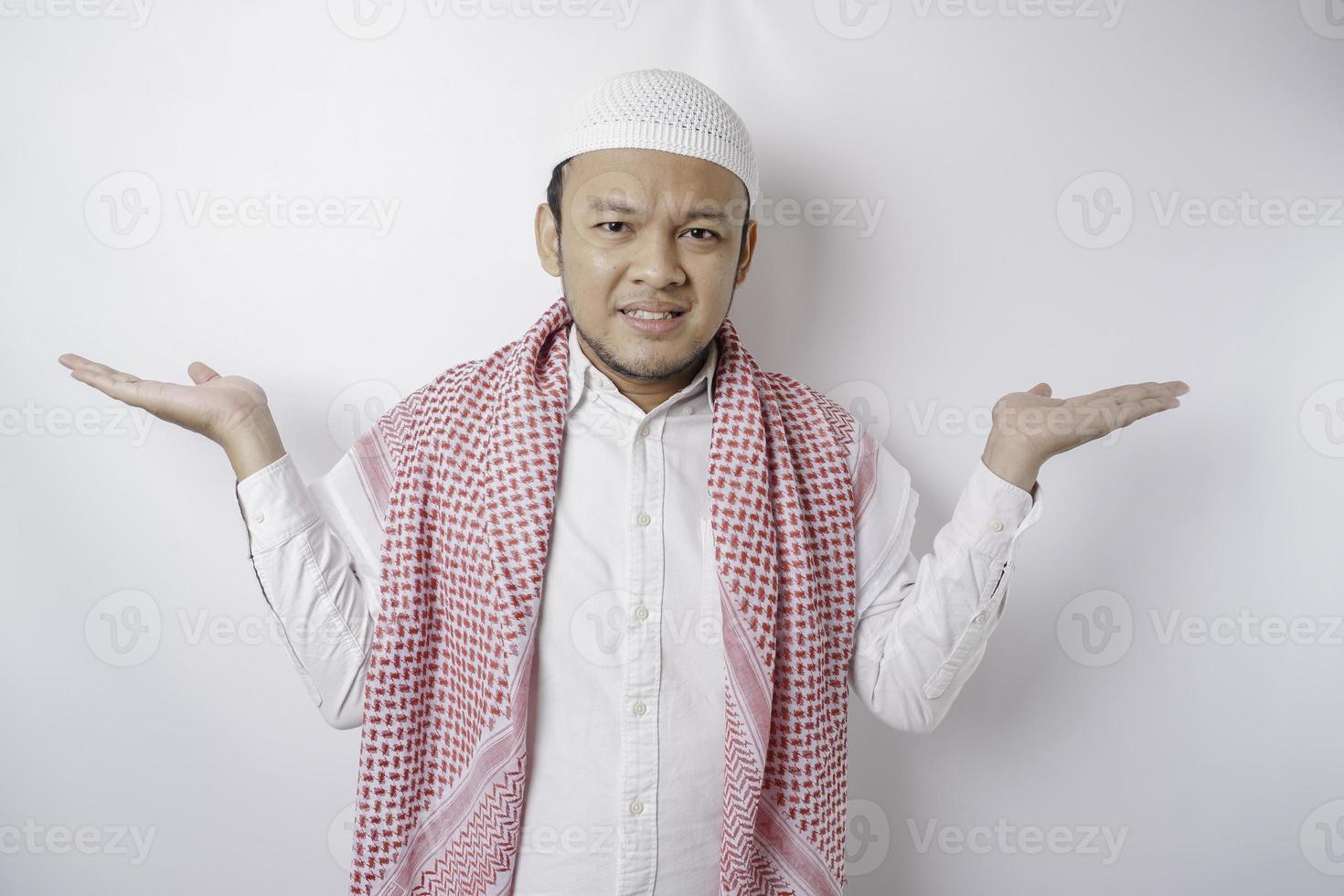 A thoughtful young Muslim man shrugging his shoulders, gesturing confusion isolated by white background photo