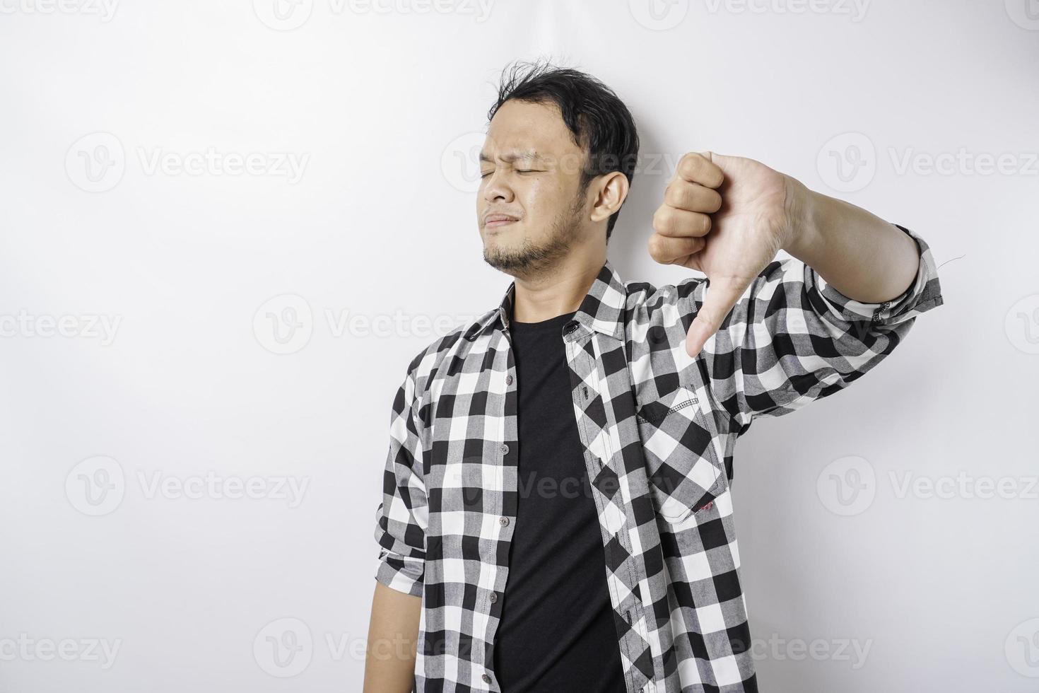 Disappointed Asian man wearing tartan short gives thumbs down hand gesture of approval, isolated by white background photo