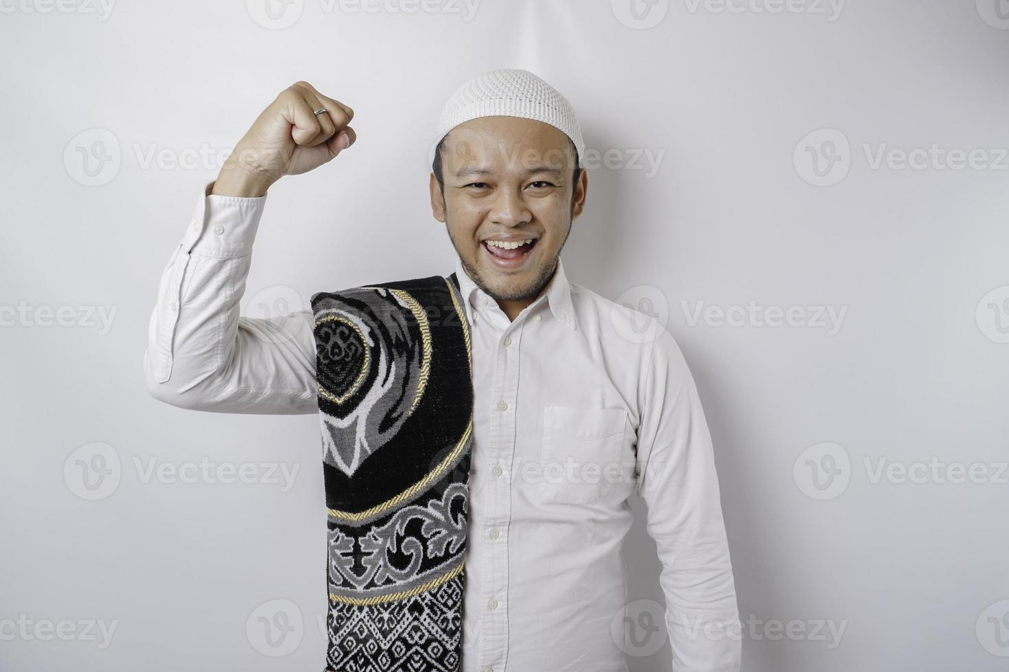 A happy young Asian Muslim man with a prayer rug on his shoulder showing a successful expression isolated by white background photo