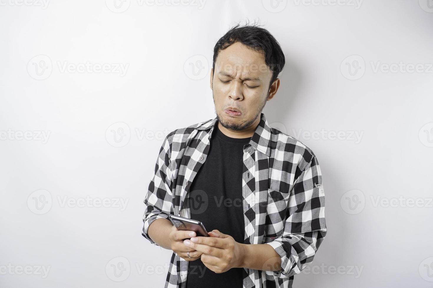 A dissatisfied young Asian man looks disgruntled wearing tartan shirt irritated face expressions holding his phone photo
