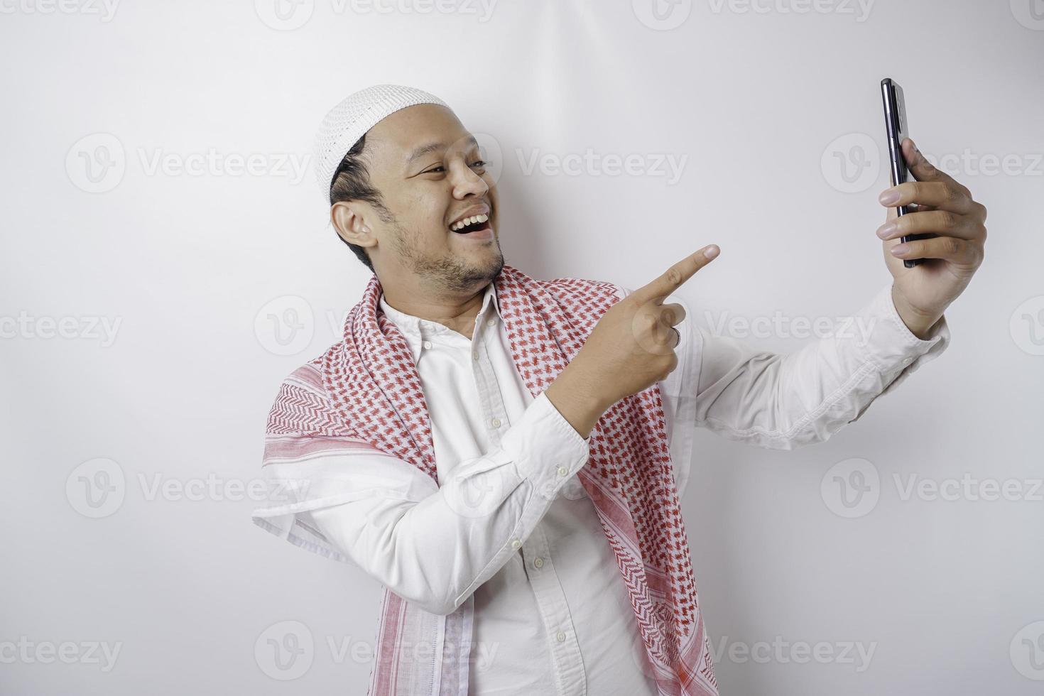 A portrait of a happy Asian Muslim man smiling while holding his phone, isolated by white background photo