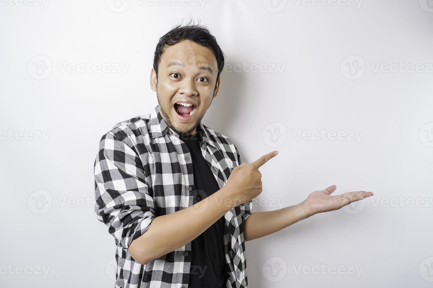 Shocked Asian man wearing tartan shirt is pointing at the copy space beside him, isolated by white background photo