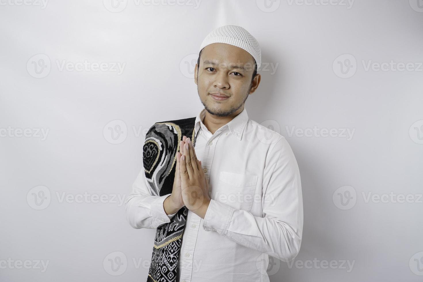 Smiling young Asian Muslim man with prayer rug on his shoulder, gesturing traditional greeting isolated over white background photo
