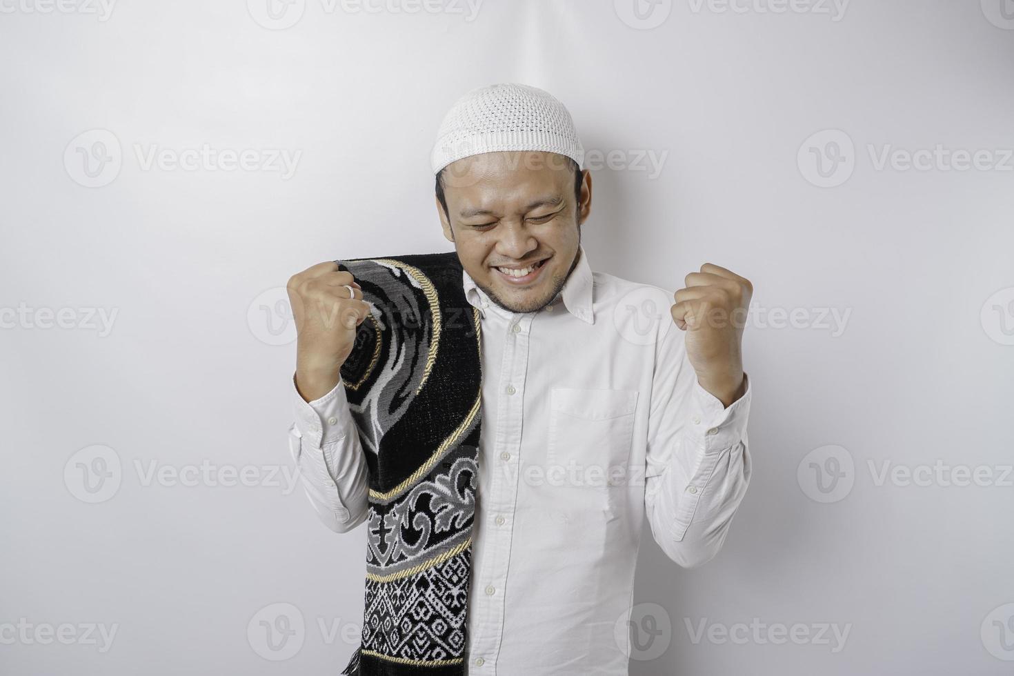 A happy young Asian Muslim man with a prayer rug on his shoulder showing a successful expression isolated by white background photo