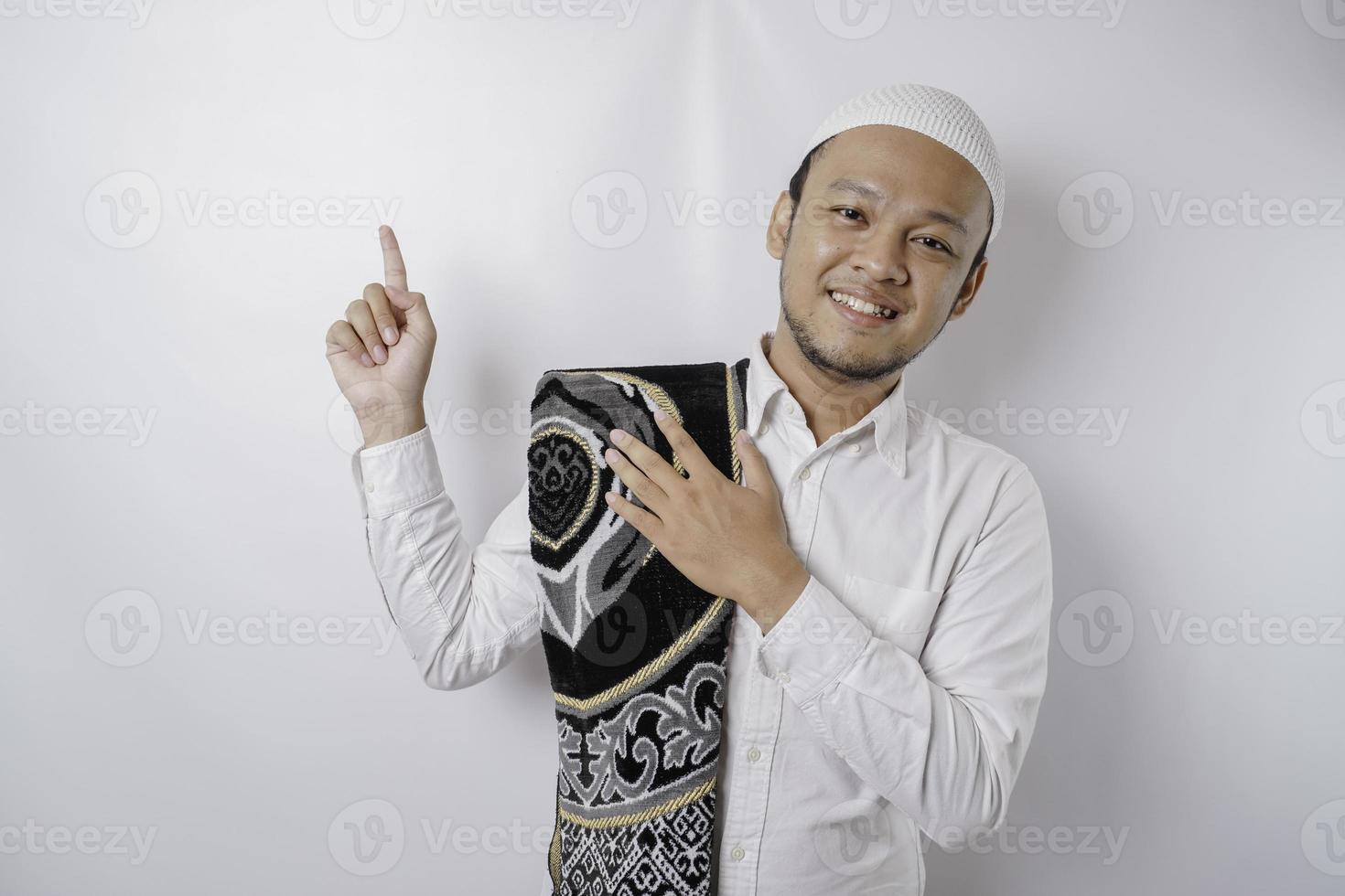 Smiling Asian Muslim man with prayer rug on his shoulder is pointing at the copy space on top of him, isolated by white background photo