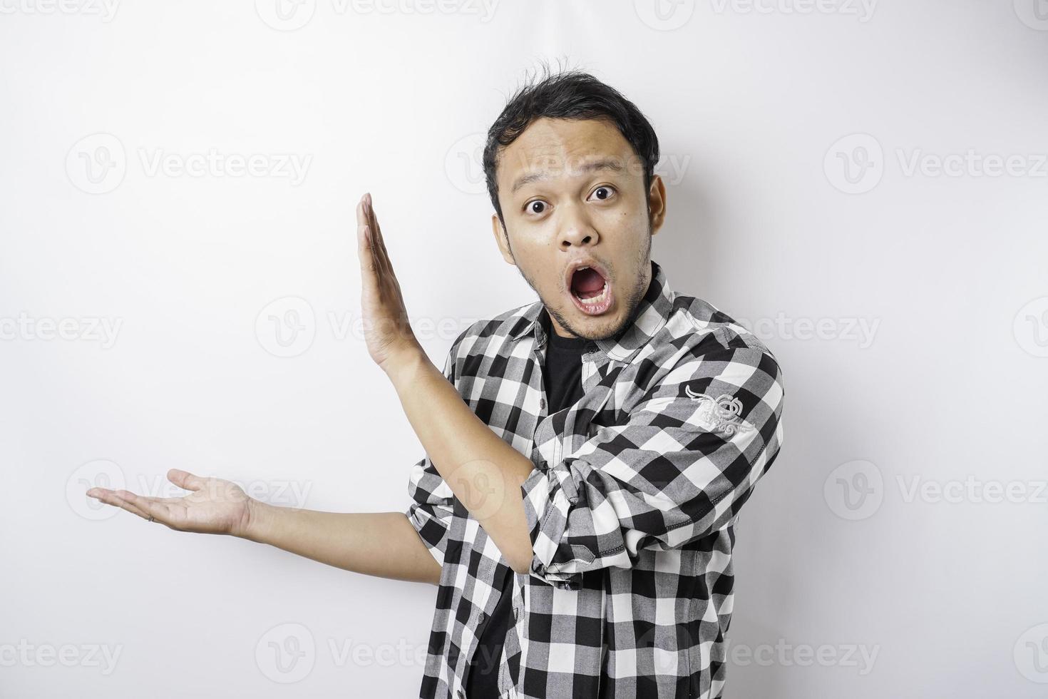 Shocked Asian man wearing tartan shirt is pointing at the copy space beside him, isolated by white background photo