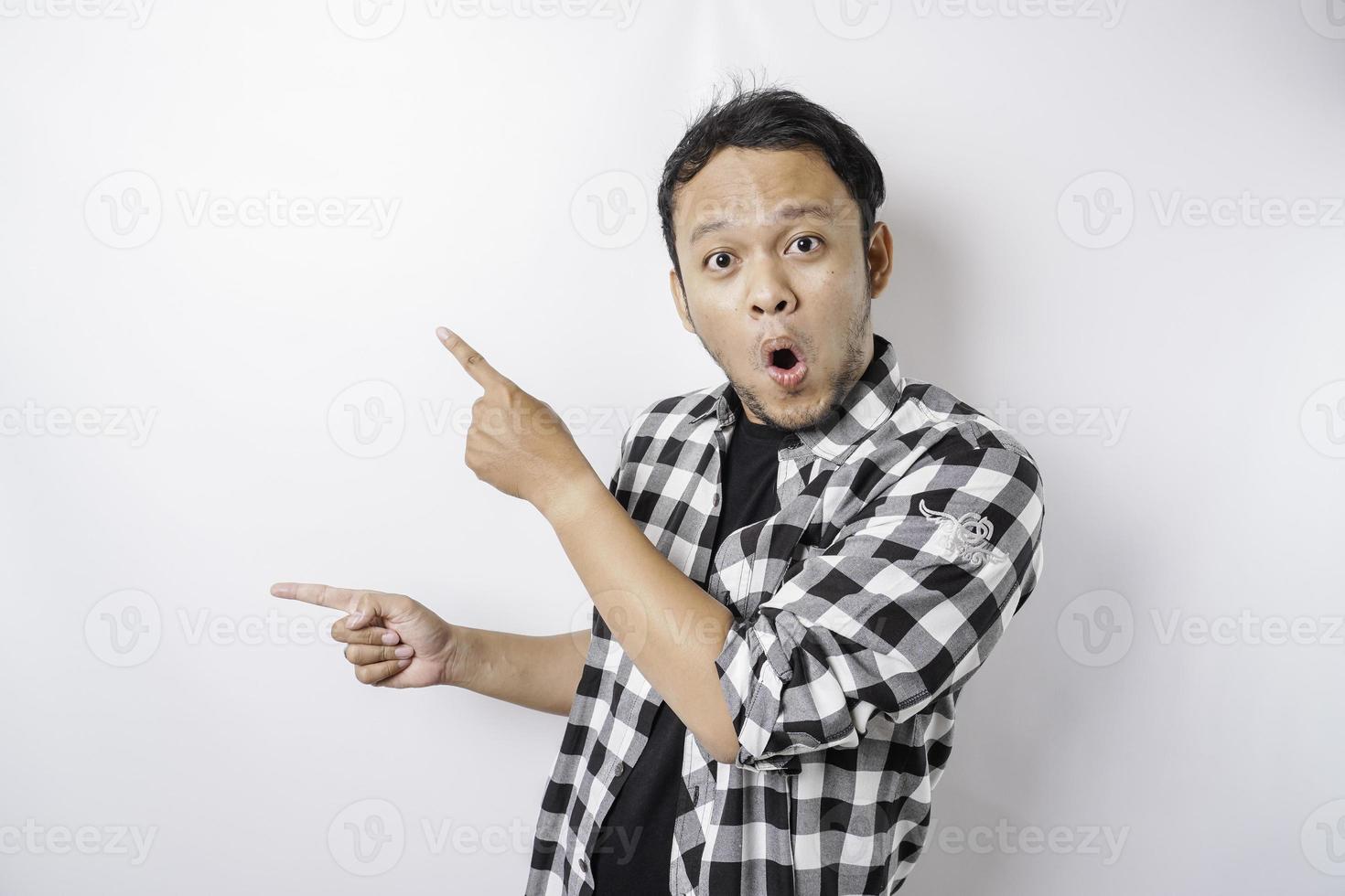 Shocked Asian man wearing tartan shirt is pointing at the copy space beside him, isolated by white background photo