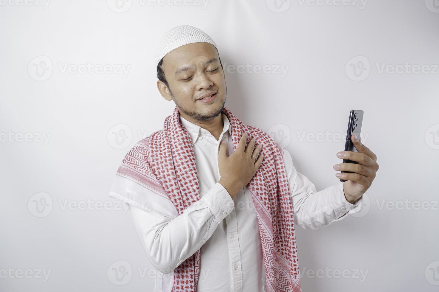 A portrait of a happy Asian Muslim man smiling while holding his phone, isolated by white background photo