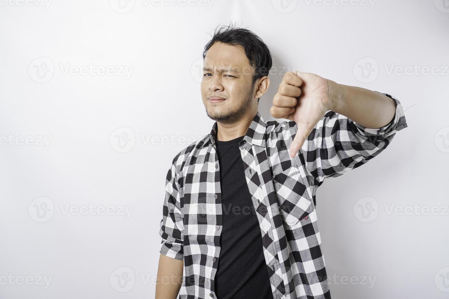 Disappointed Asian man wearing tartan short gives thumbs down hand gesture of approval, isolated by white background photo