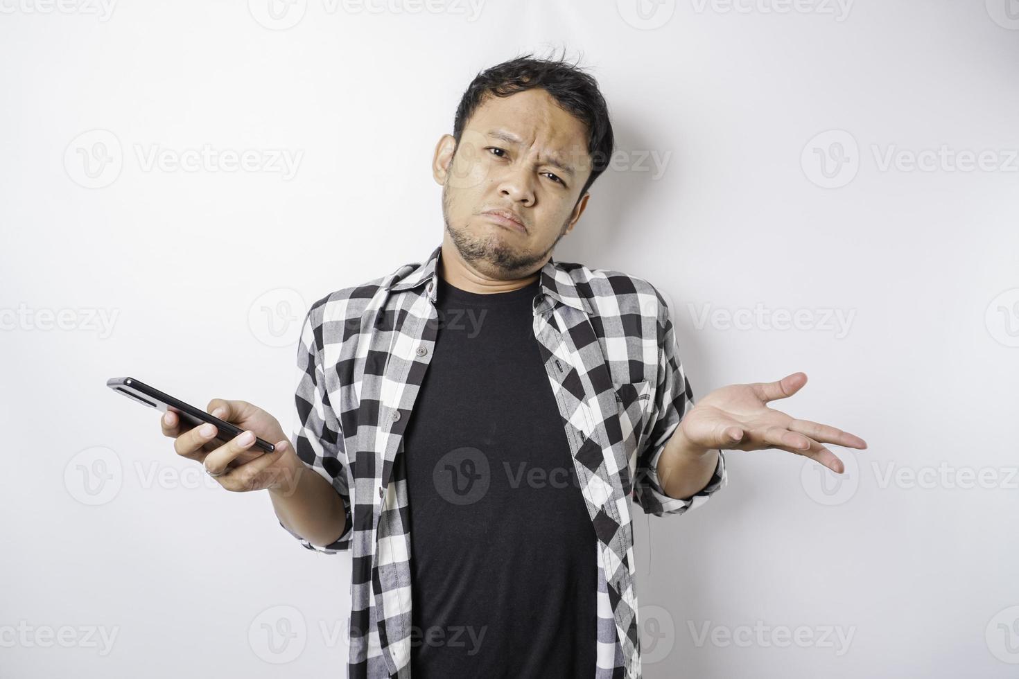 A dissatisfied young Asian man looks disgruntled wearing tartan shirt irritated face expressions holding his phone photo