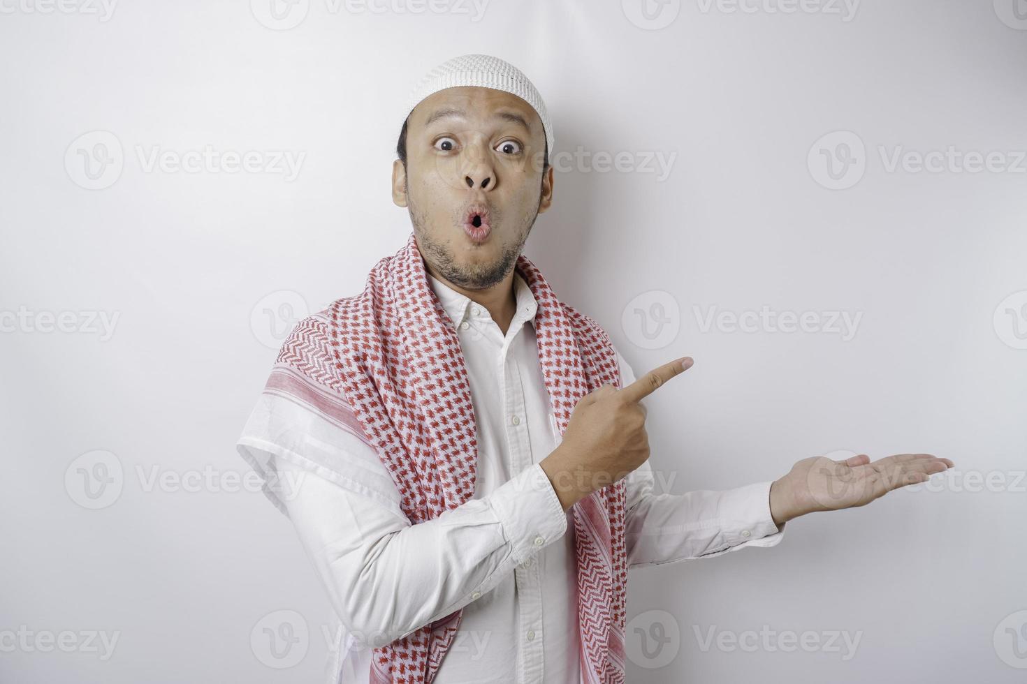 Shocked Asian Muslim man pointing at the copy space beside him, isolated by white background photo